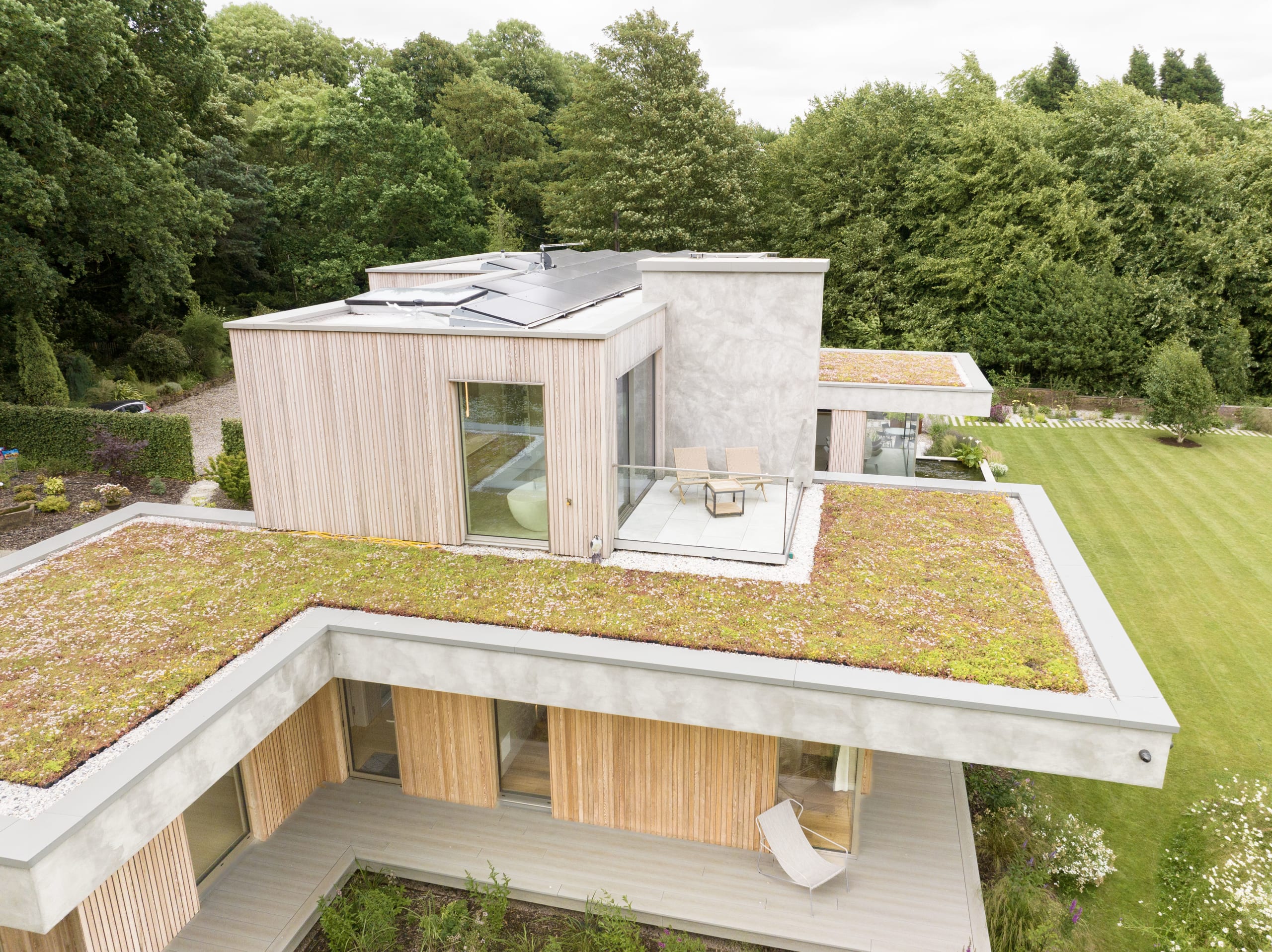 Aerial view of a modern house with sedum green roofs, timber cladding, rooftop terrace and solar panels amid lawn and trees