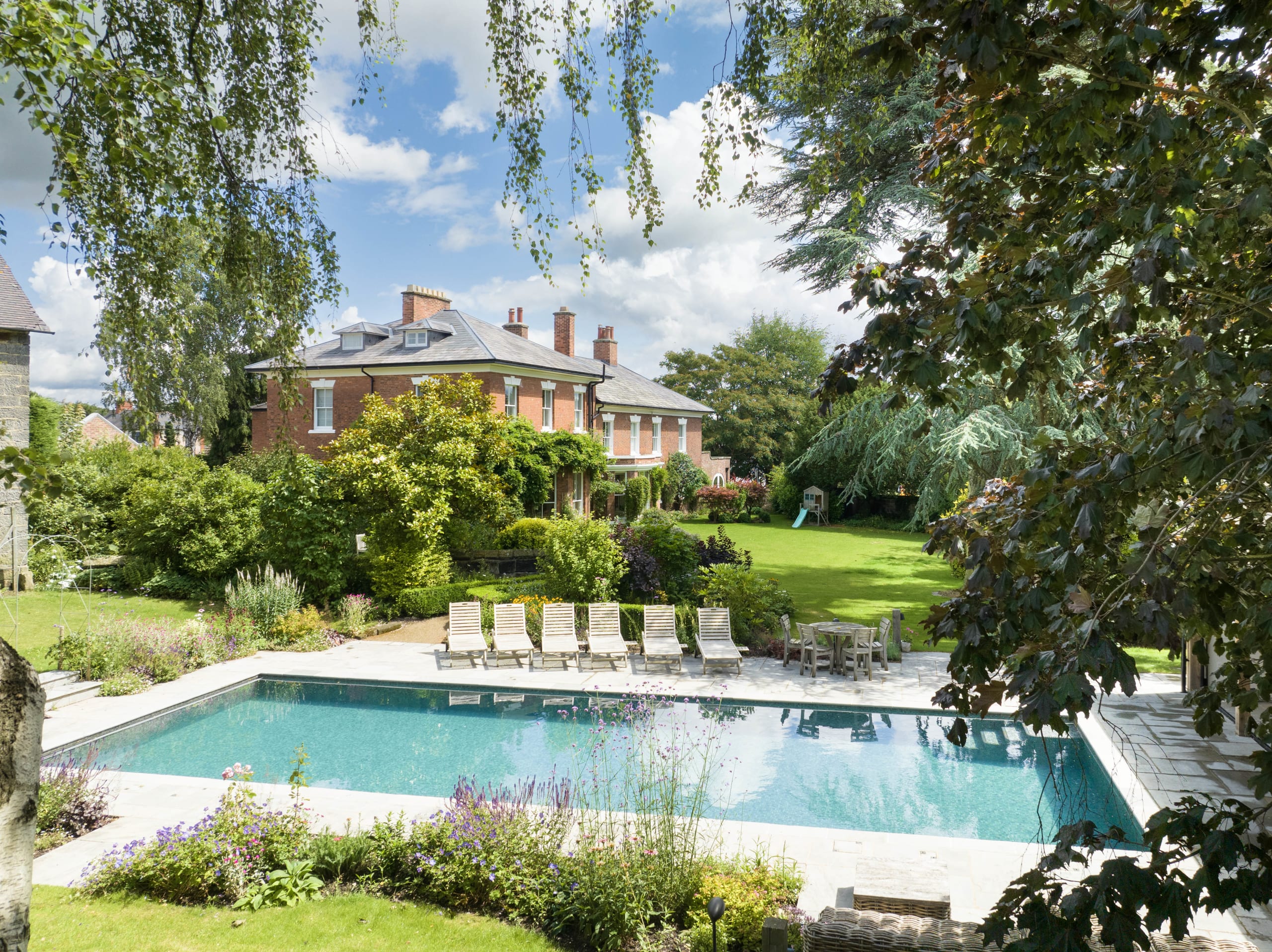 Georgian red brick manor house with slate roof overlooking manicured lawn, swimming pool, and landscaped garden terrace