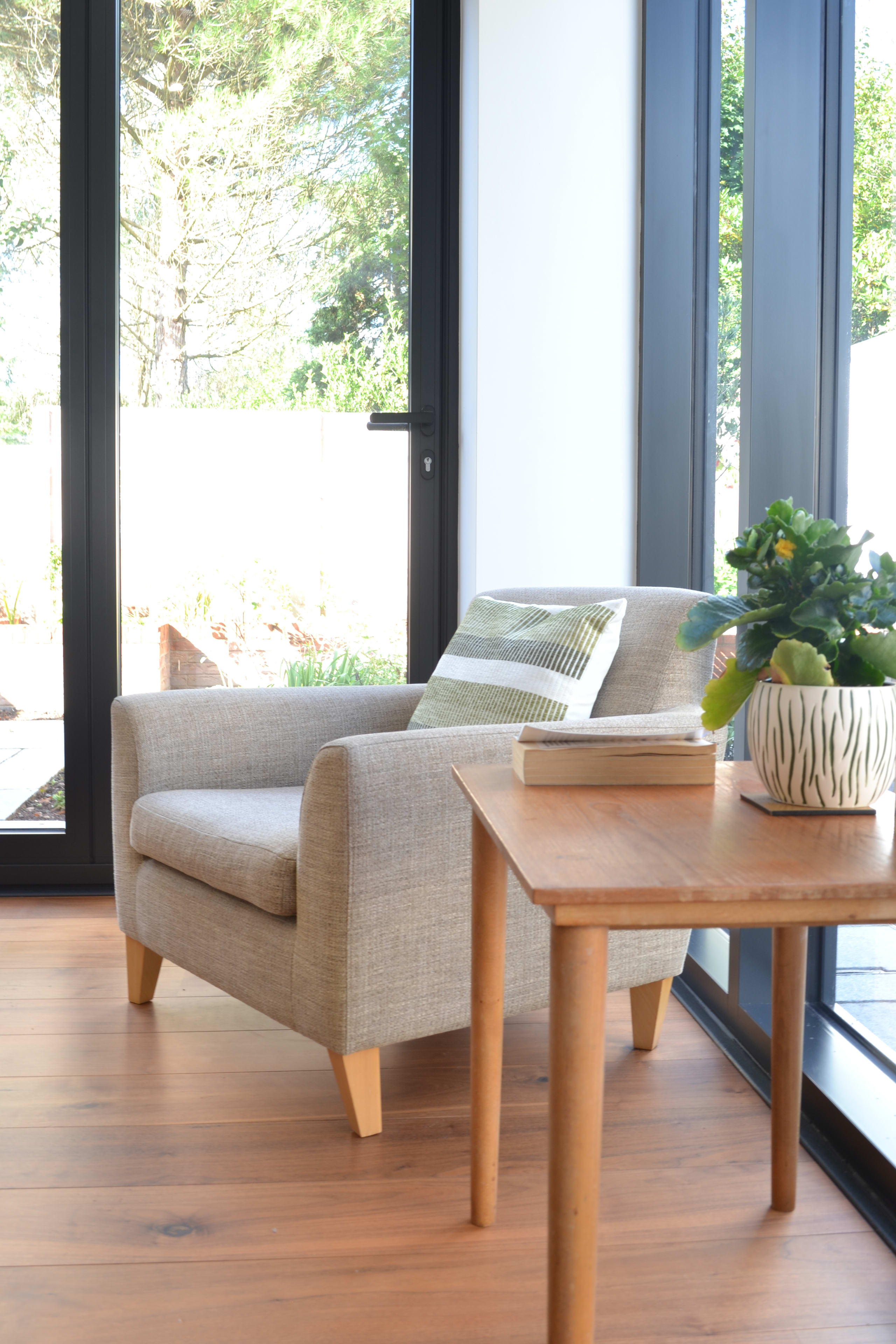 Modern reading nook with gray upholstered armchair, wooden side table with plant, and floor-to-ceiling black-framed windows