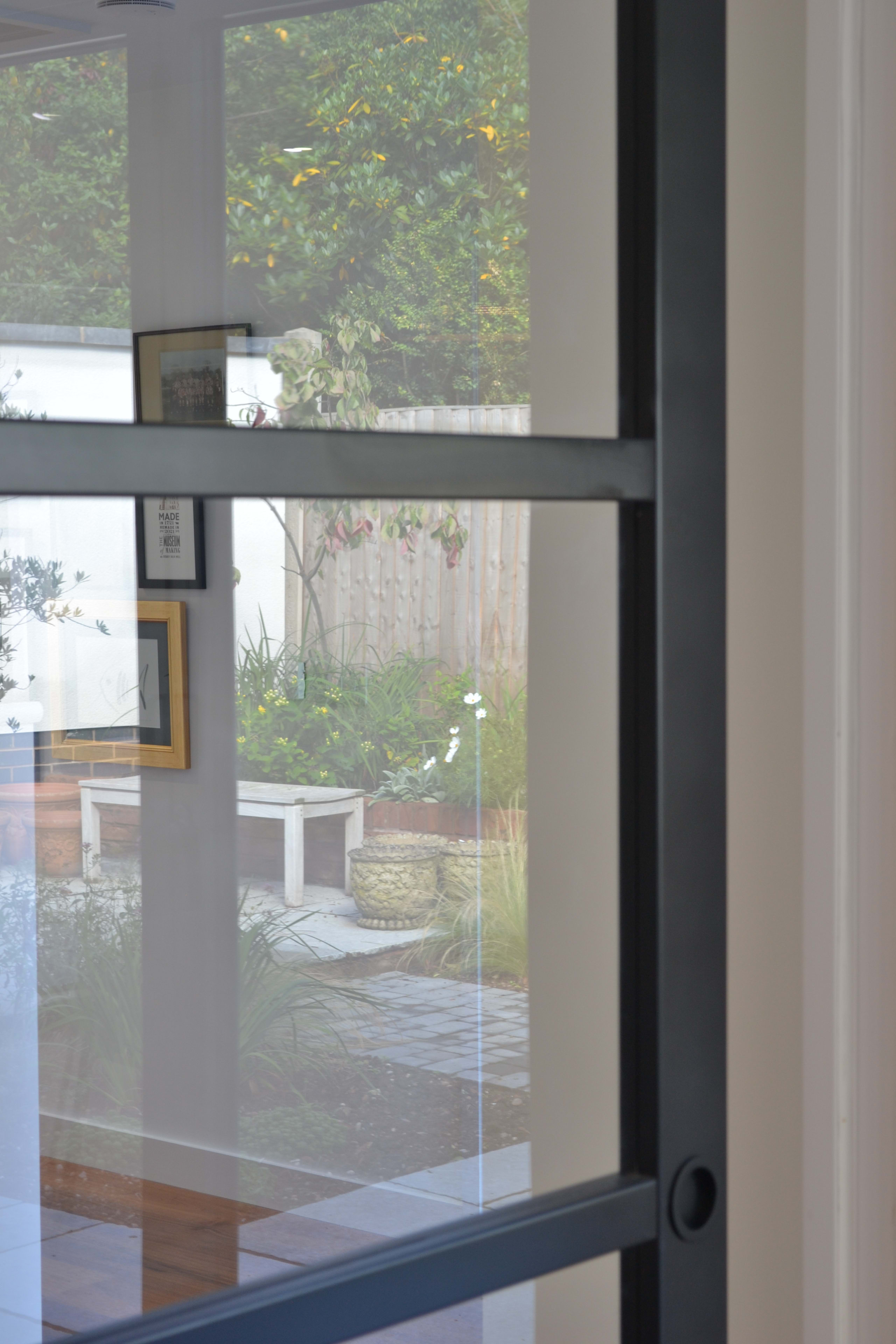 Black steel-framed glass door looking out to courtyard garden with potted plants, bench, and wooden fence