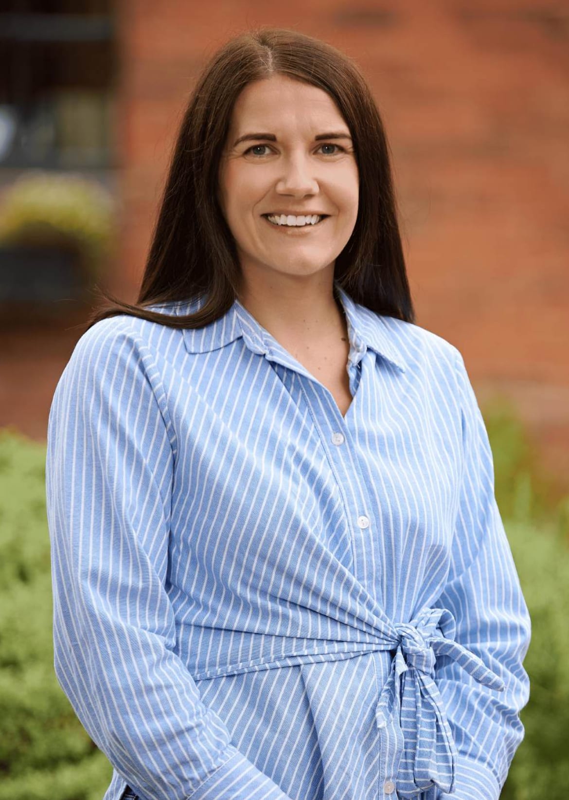 Woman with long brown hair smiling outdoors in a blue-and-white striped shirt, brick building behind her