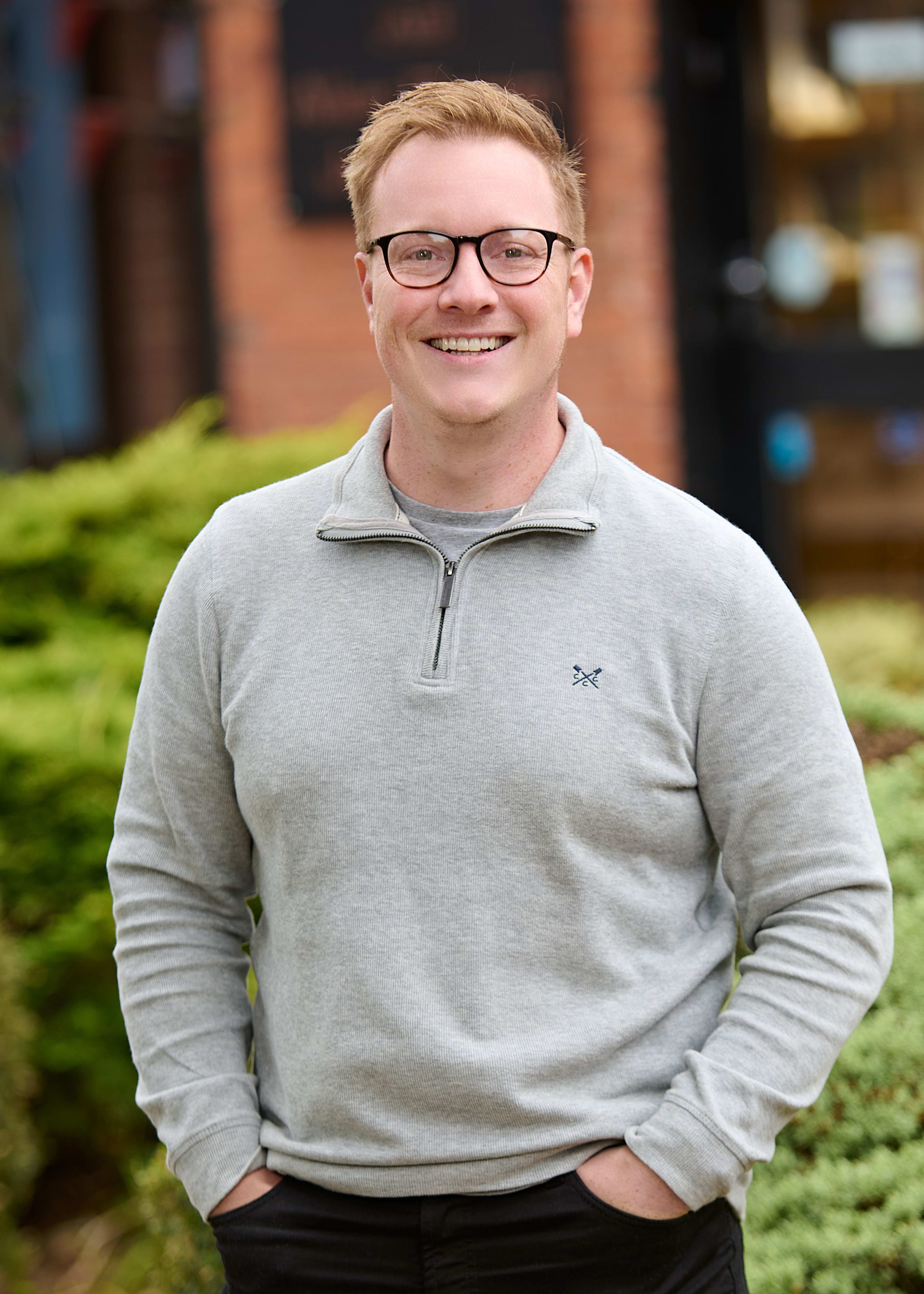 Smiling man with red hair and glasses wearing a gray quarter-zip sweater, standing outdoors with hands in pockets