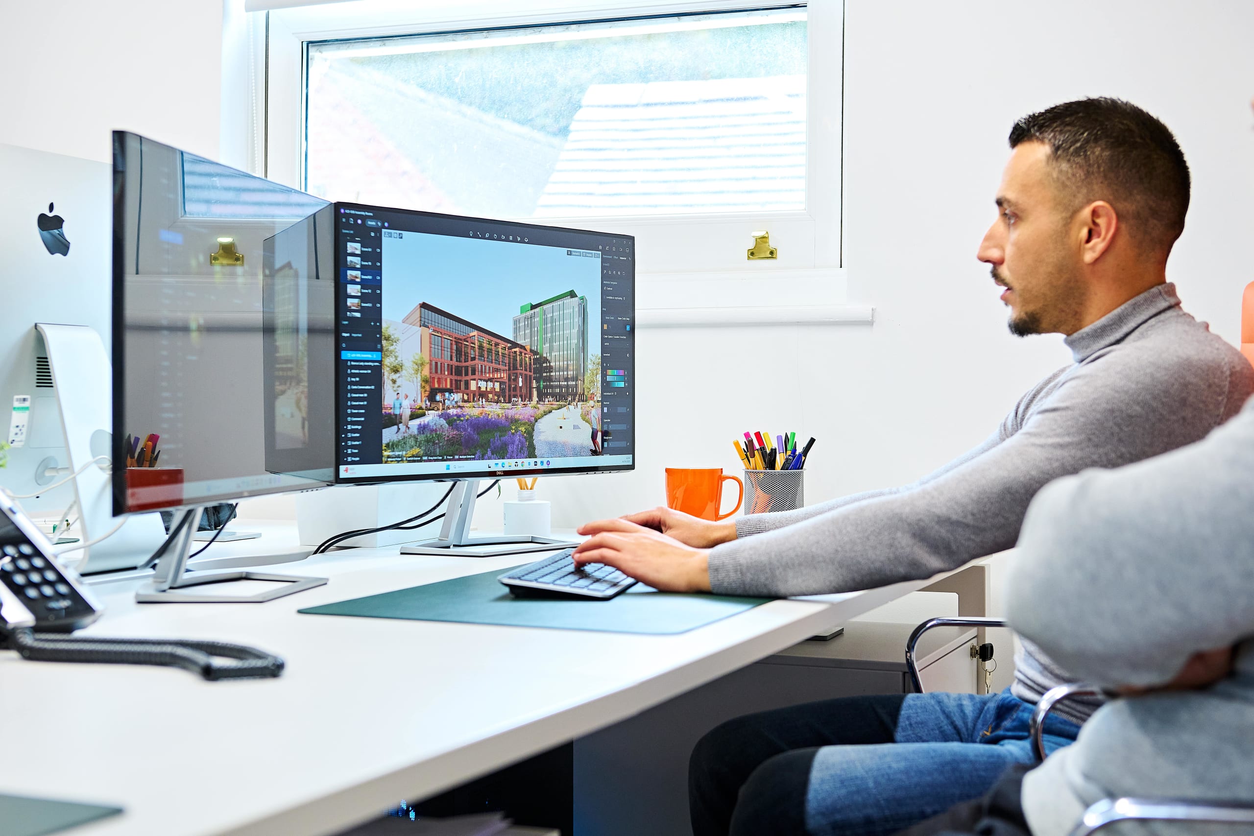 Architect working at desk with dual monitors displaying 3D rendering of mixed-use urban development project