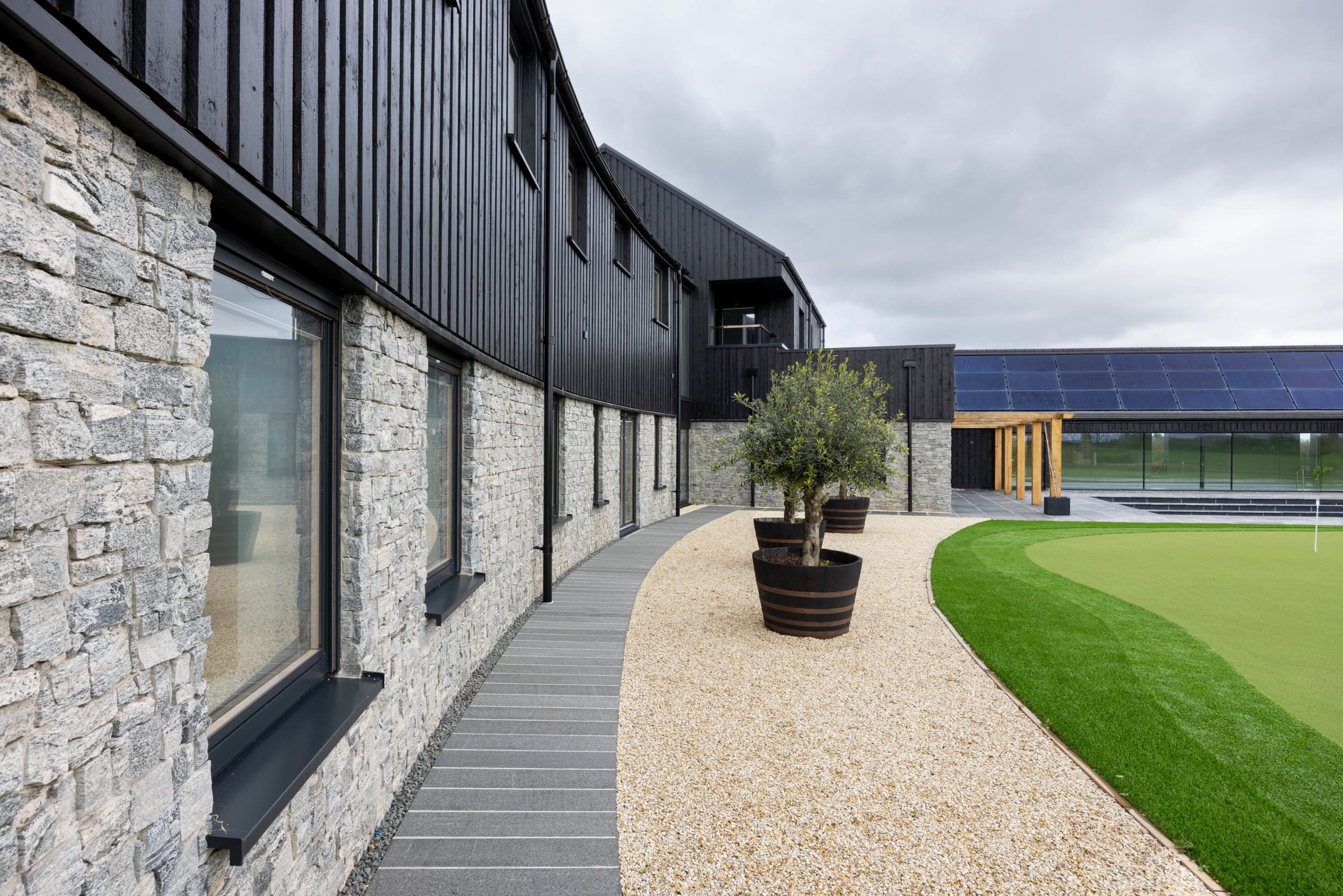 Curved stone and black timber-clad building beside a gravel path and putting green, with solar panels visible beyond