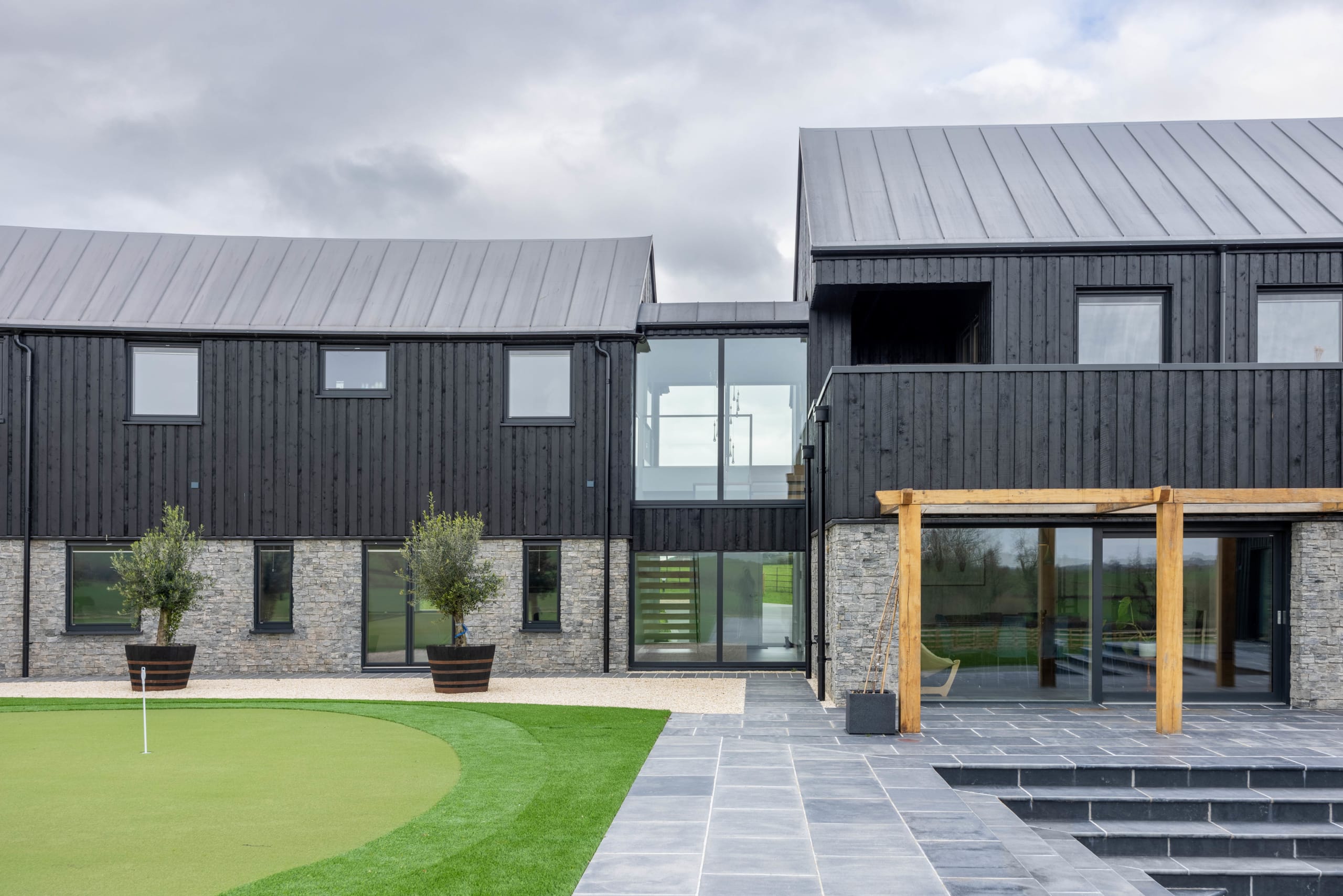Black timber-clad barn-style house with stone base, glass gable, and timber pergola beside a putting green