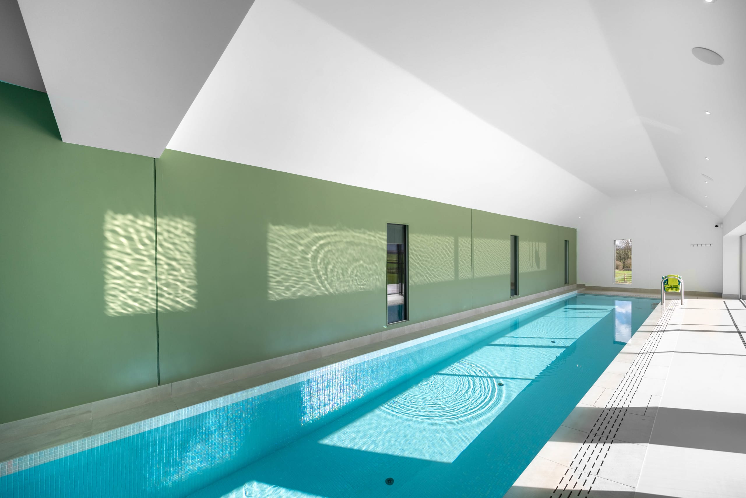Indoor lap pool with sage-green walls reflecting rippling water light beneath a vaulted white ceiling