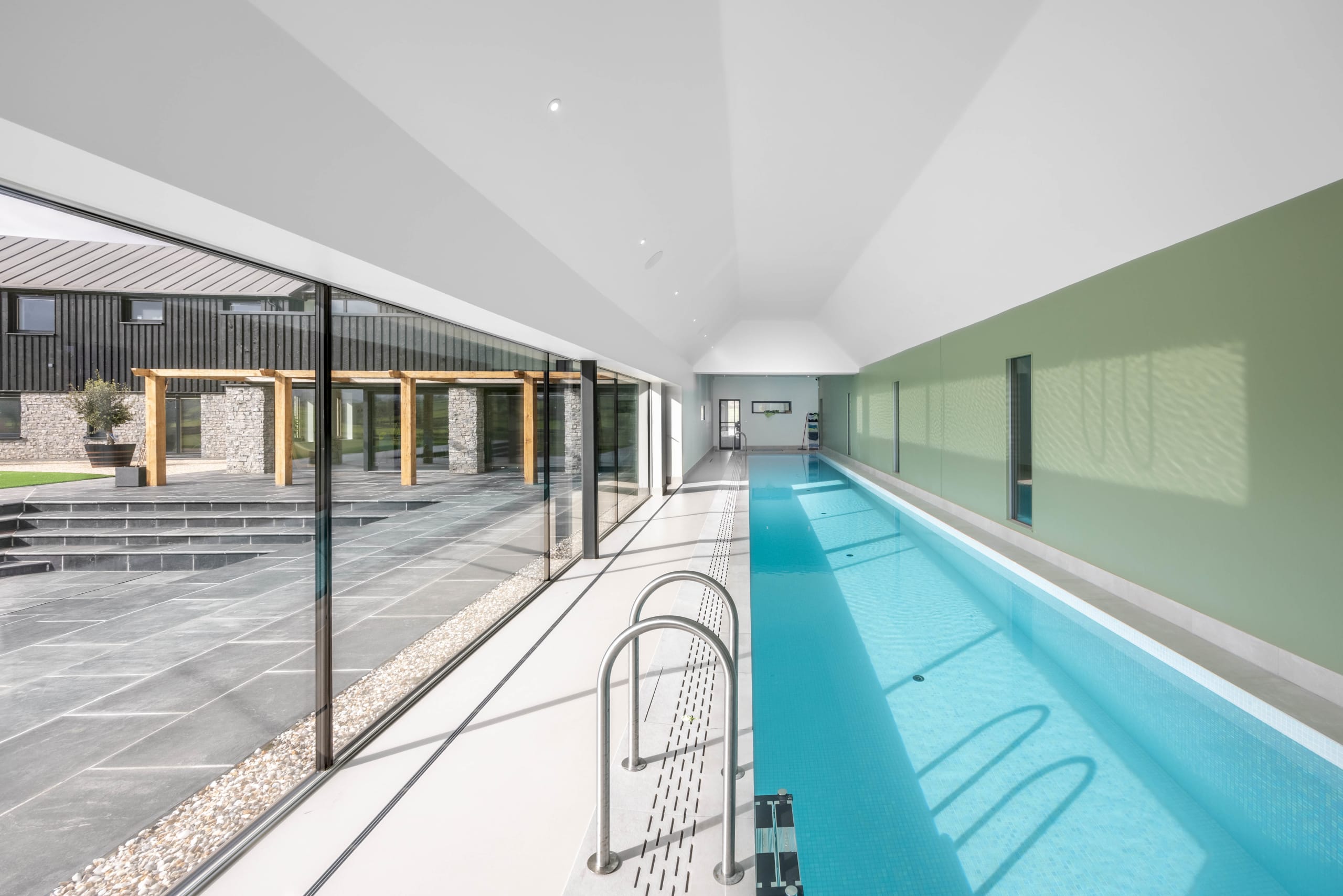 Indoor lap pool with vaulted white ceiling, green feature wall, and glazed wall overlooking a stone-paved courtyard