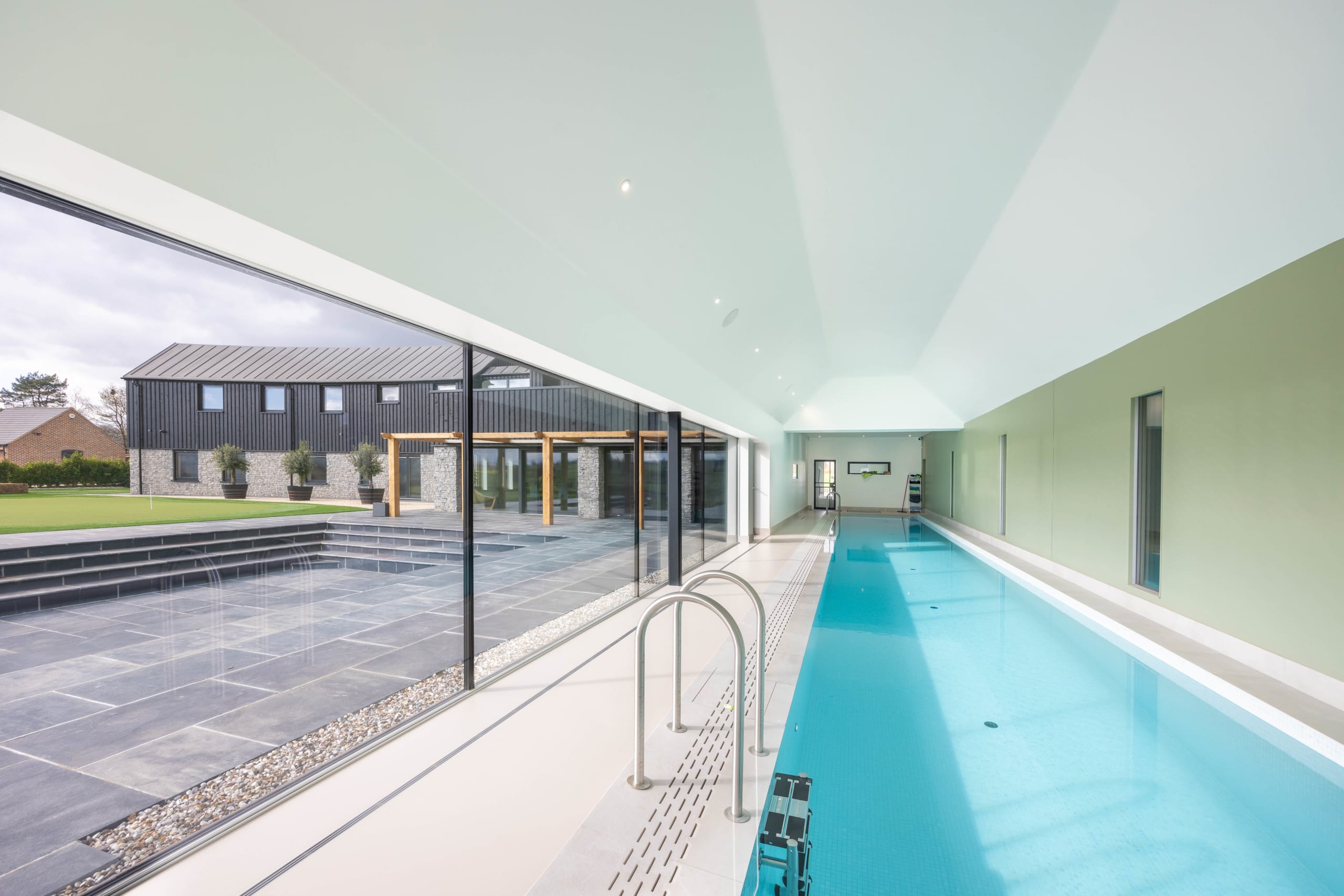 Long indoor swimming pool with glazed wall overlooking a stone terrace and dark timber-clad barn conversion