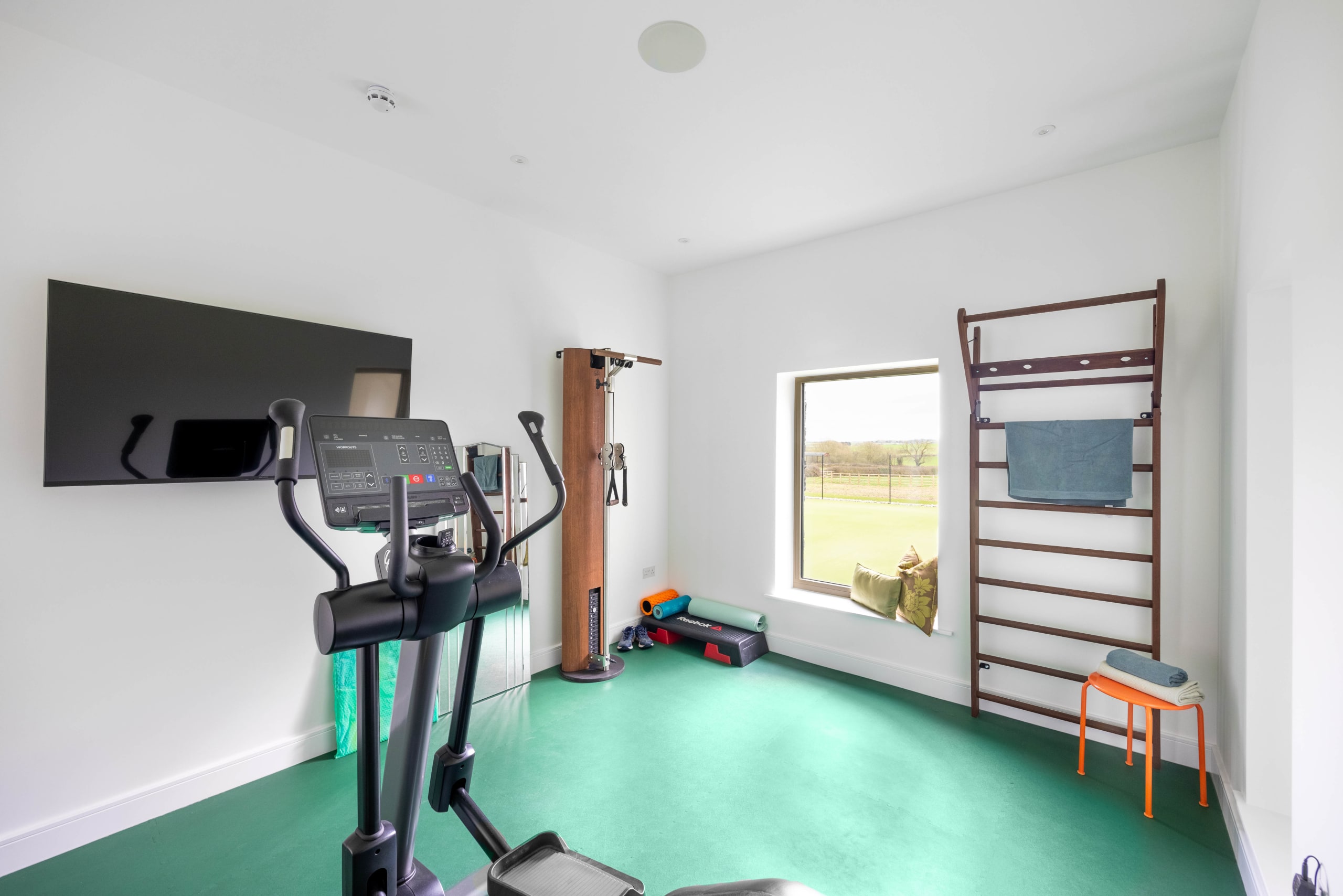 Home gym with green rubber flooring, wall-mounted TV, cross trainer, and wooden wall bars beside a window overlooking fields