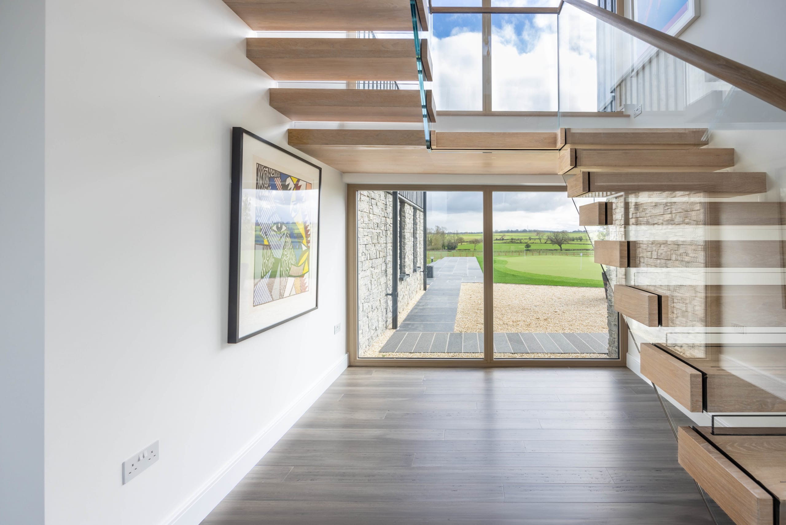 Cantilevered timber staircase with glass balustrade beside a hallway framed in stone leading to countryside views
