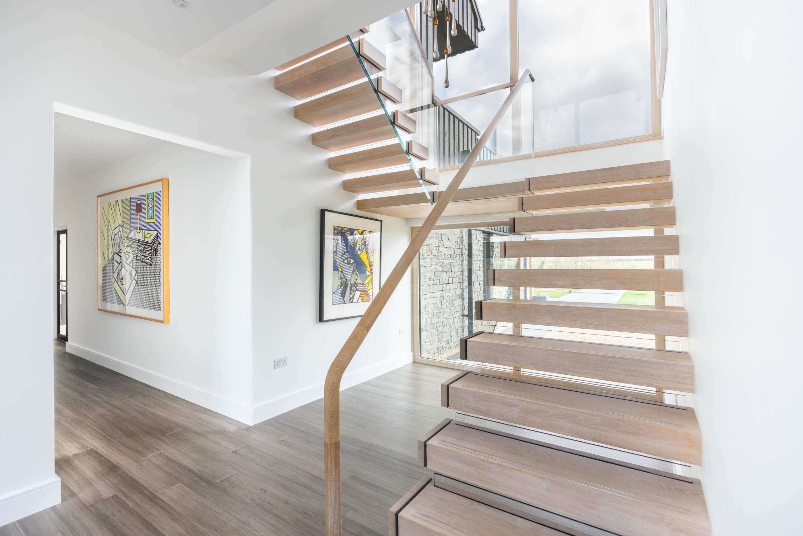 Floating timber staircase with glass balustrade and wood handrail beside a white double-height entrance hall