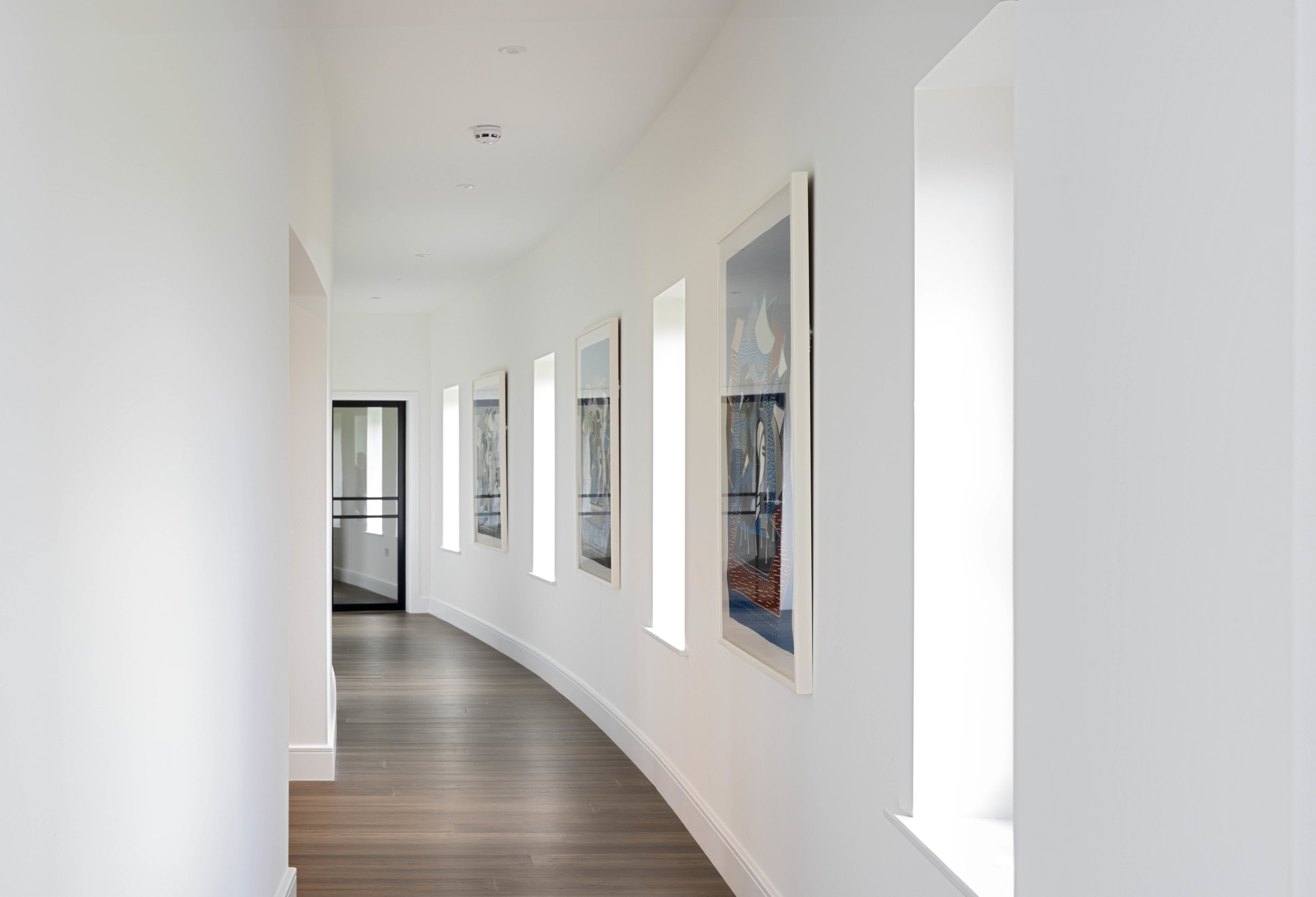 Curved white corridor with narrow vertical windows and framed artwork above dark timber flooring