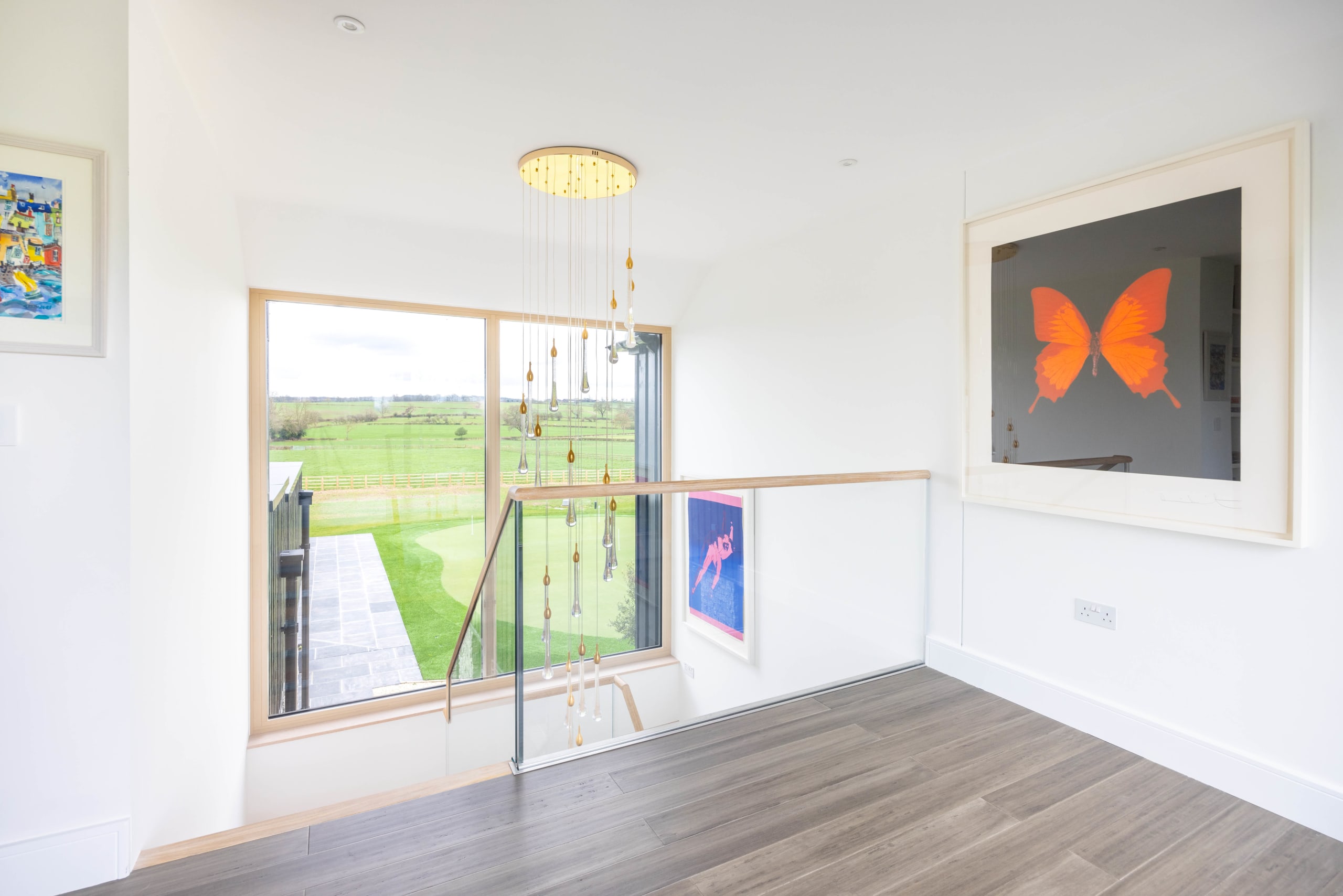 Landing with glass-balustrade staircase, timber floor, large window over green fields, and an orange butterfly artwork