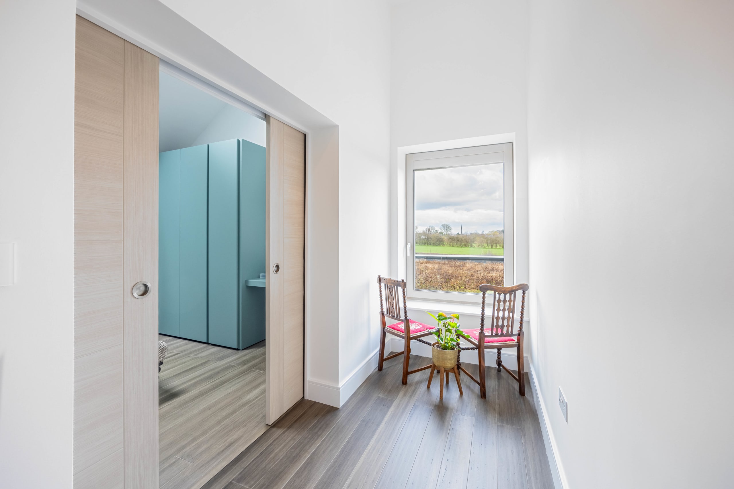 Hallway with sliding oak doors opening to a teal-cabineted bathroom, beside a window seat with two vintage chairs