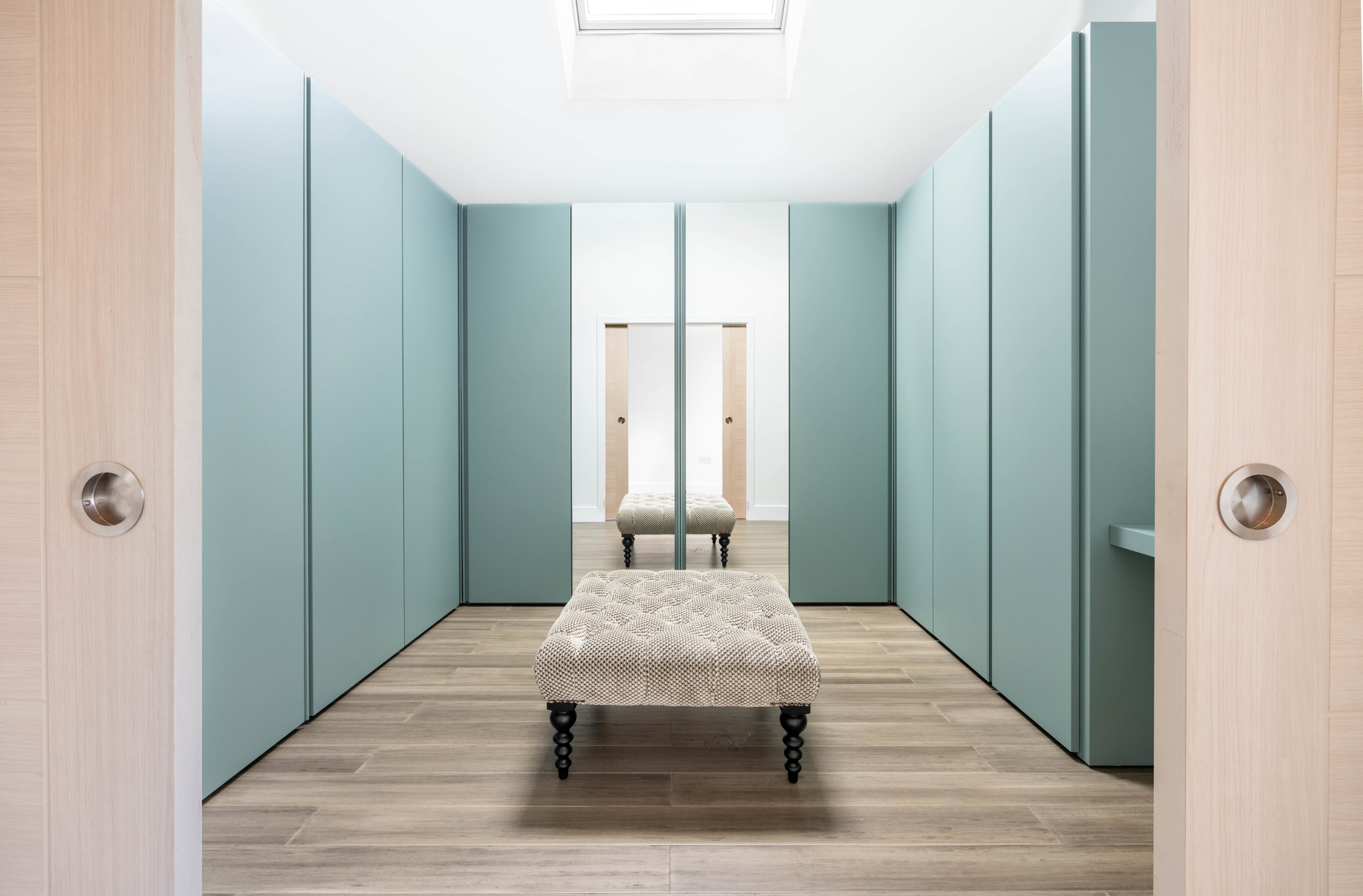 Walk-in dressing room with sage-green floor-to-ceiling wardrobes and a tufted bench beneath a skylight