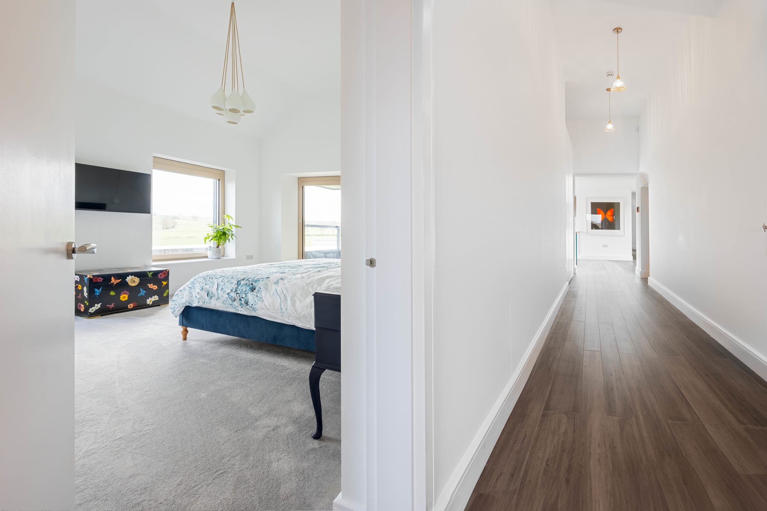 White hallway with dark timber flooring leading past a bedroom with blue upholstered bed and countryside view