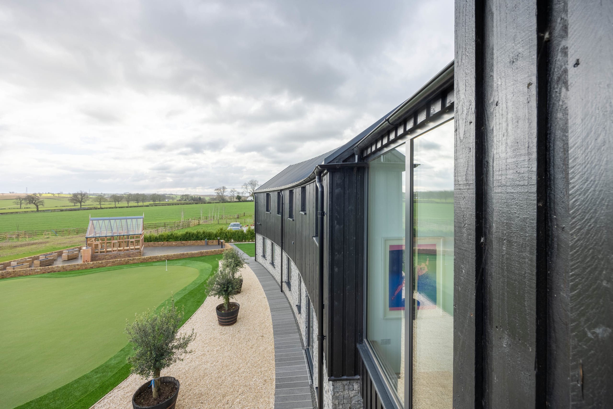 Curved black timber-clad building overlooking a putting green, with potted olive trees and gravel terrace