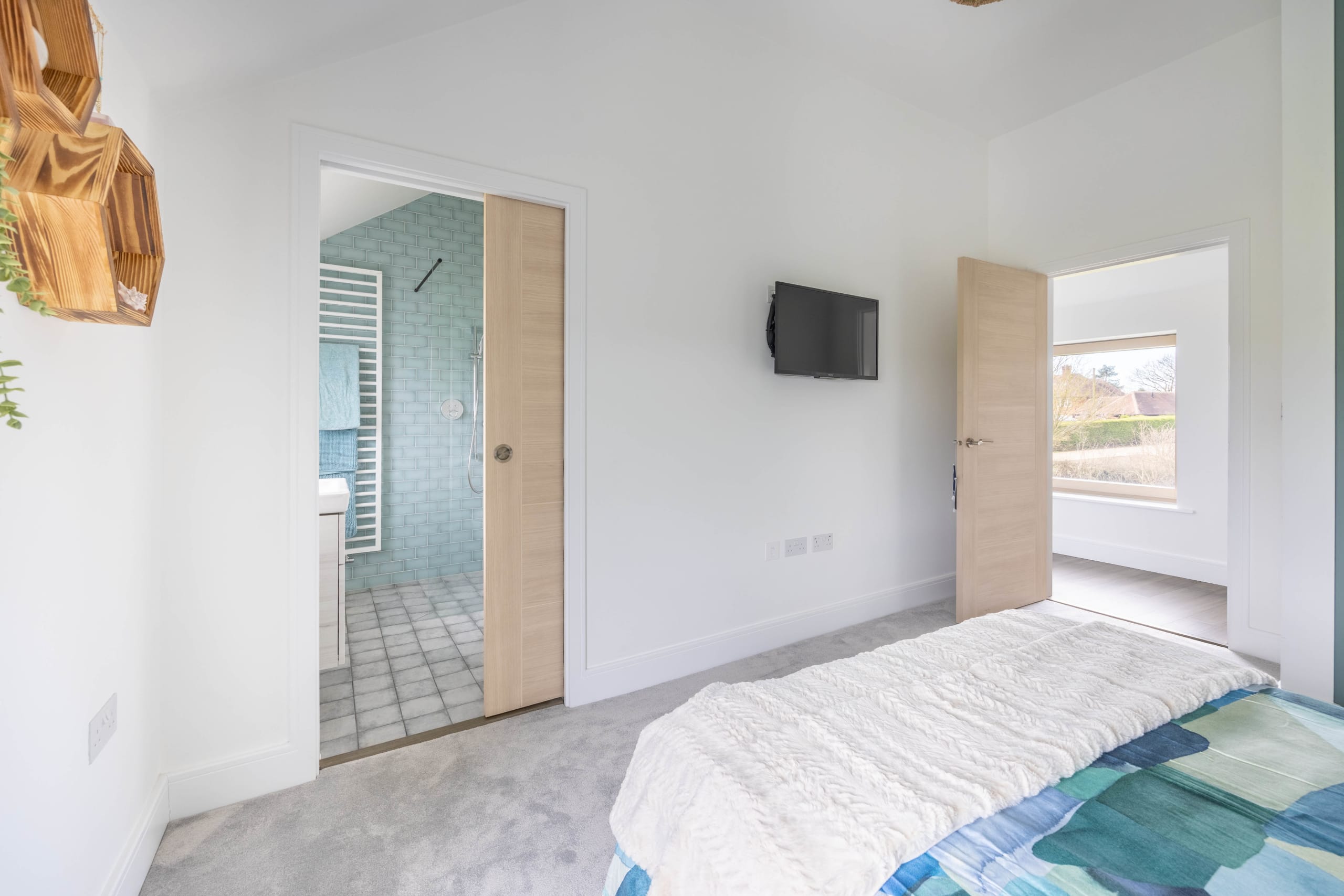 Bright bedroom with wall-mounted TV, en-suite bathroom with teal tiled shower, and open door to hallway with window