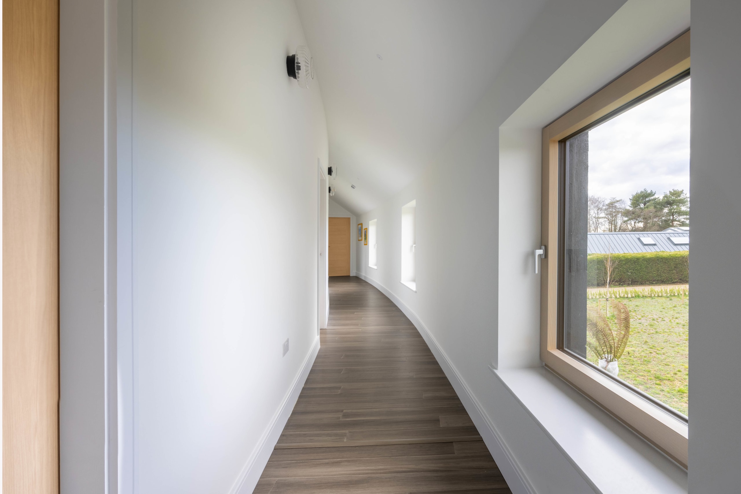 Curved white hallway with dark wood flooring and a large timber-framed window overlooking a garden