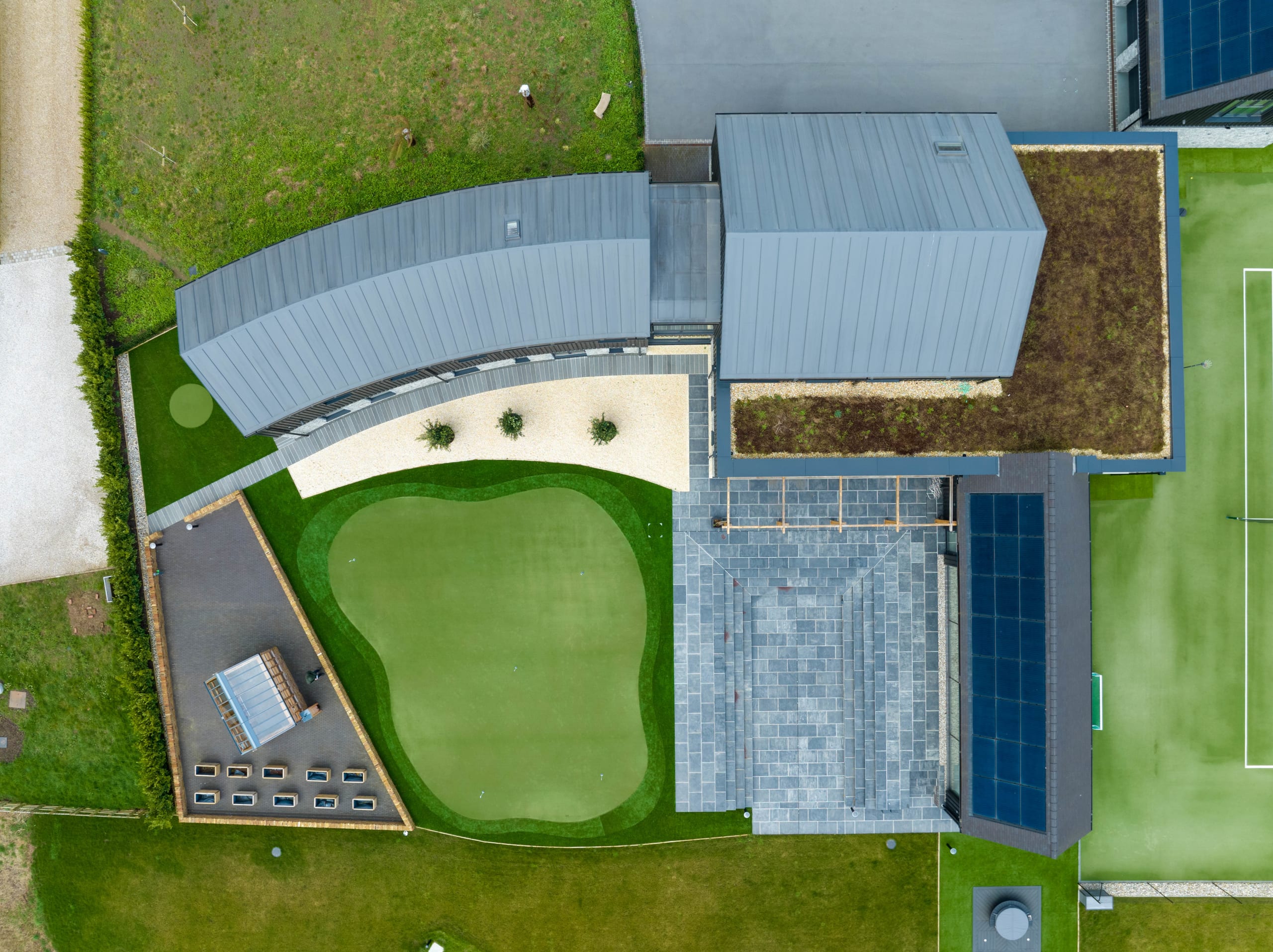 Aerial view of a curved standing-seam metal roof building with green roof and adjacent gravel courtyard and lawn