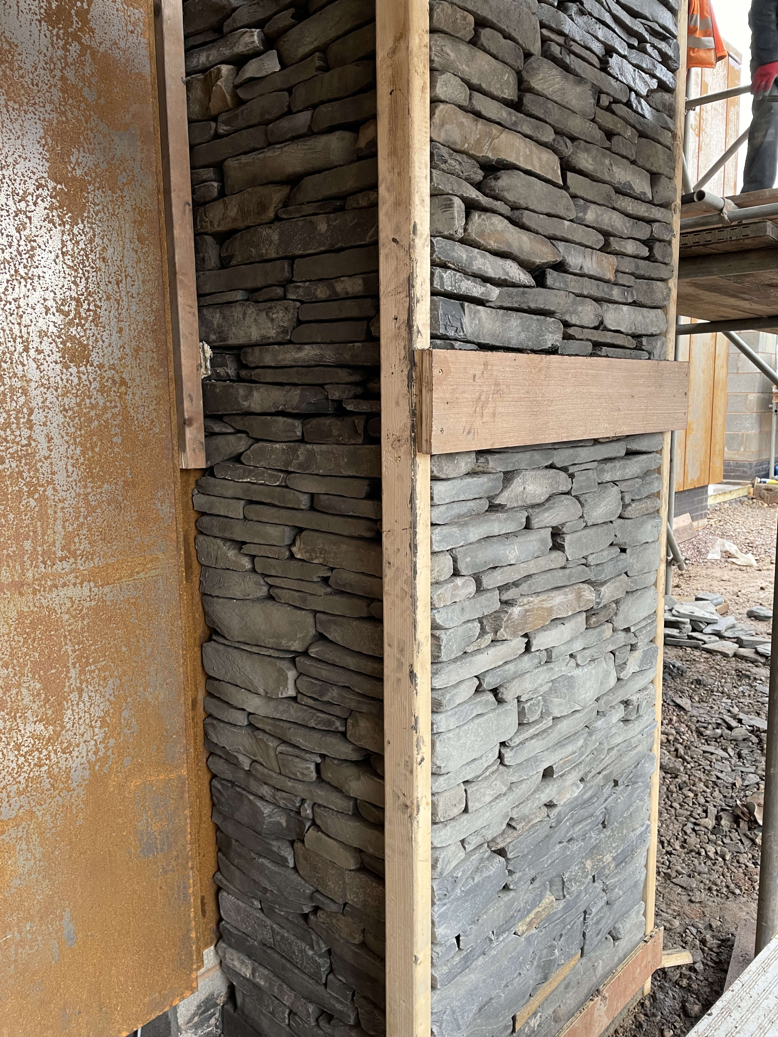 Dry stone wall construction with stacked slate stones and timber frame supports at building corner on active construction site