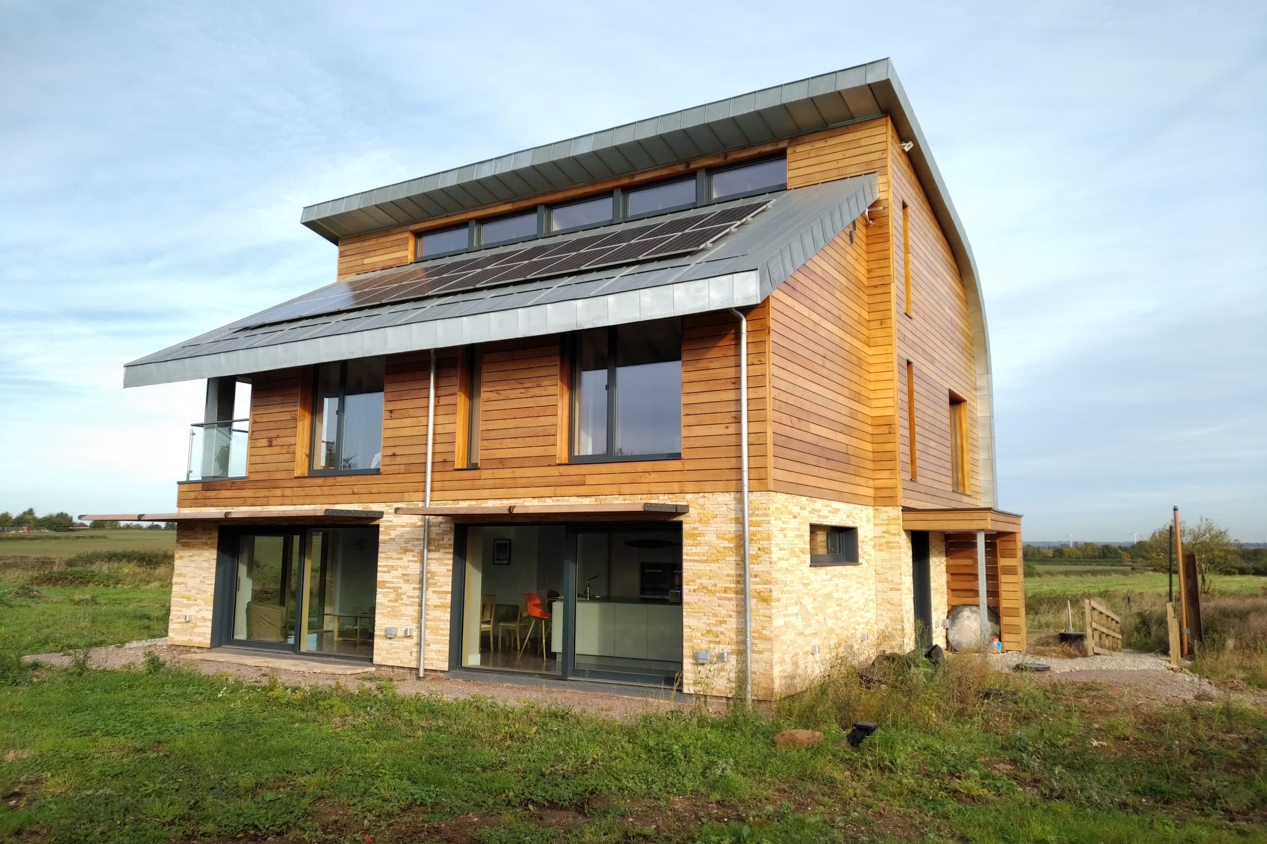 Modern two-story eco home with cedar cladding, stone base, curved zinc roof, and solar panels in rural countryside setting