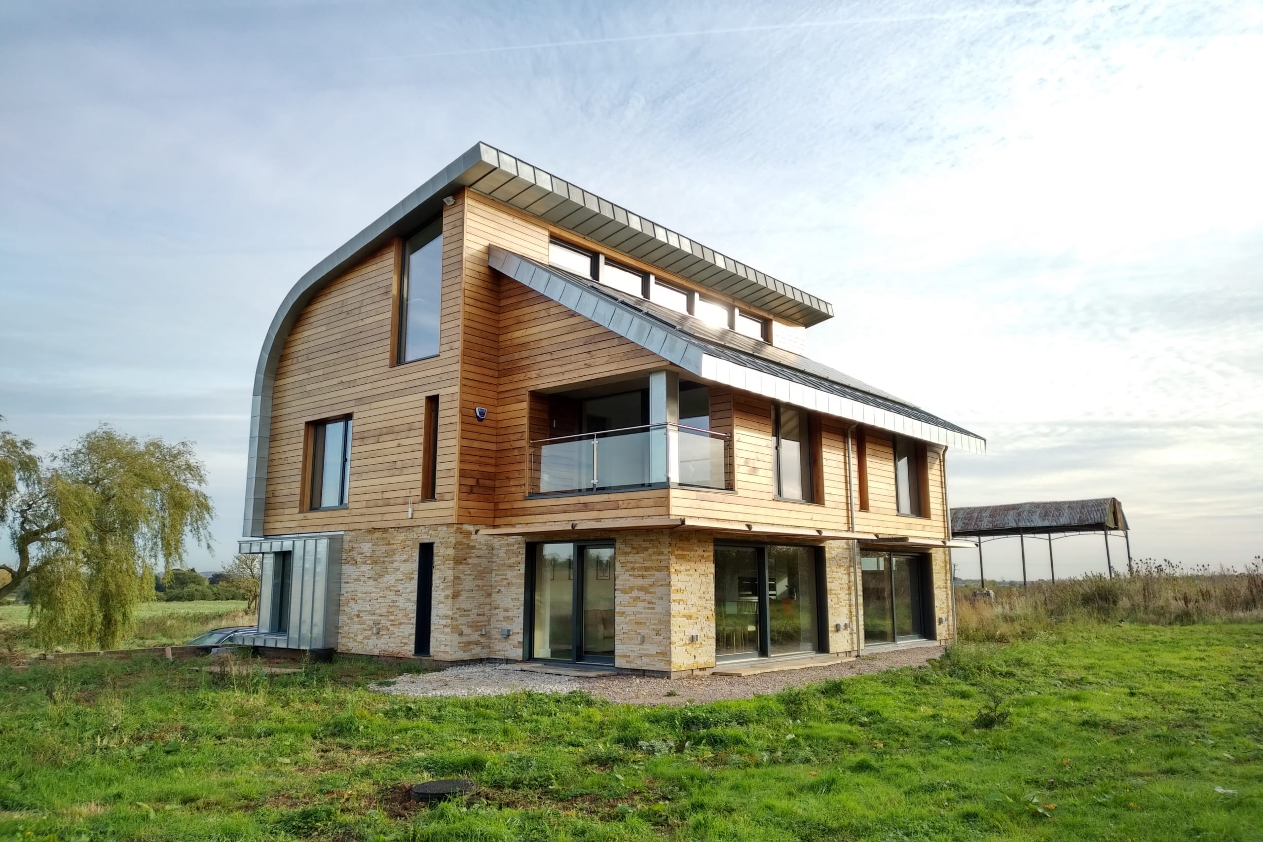 Modern two-story rural home with timber cladding, stone base, curved zinc roof, and large glass windows in open countryside