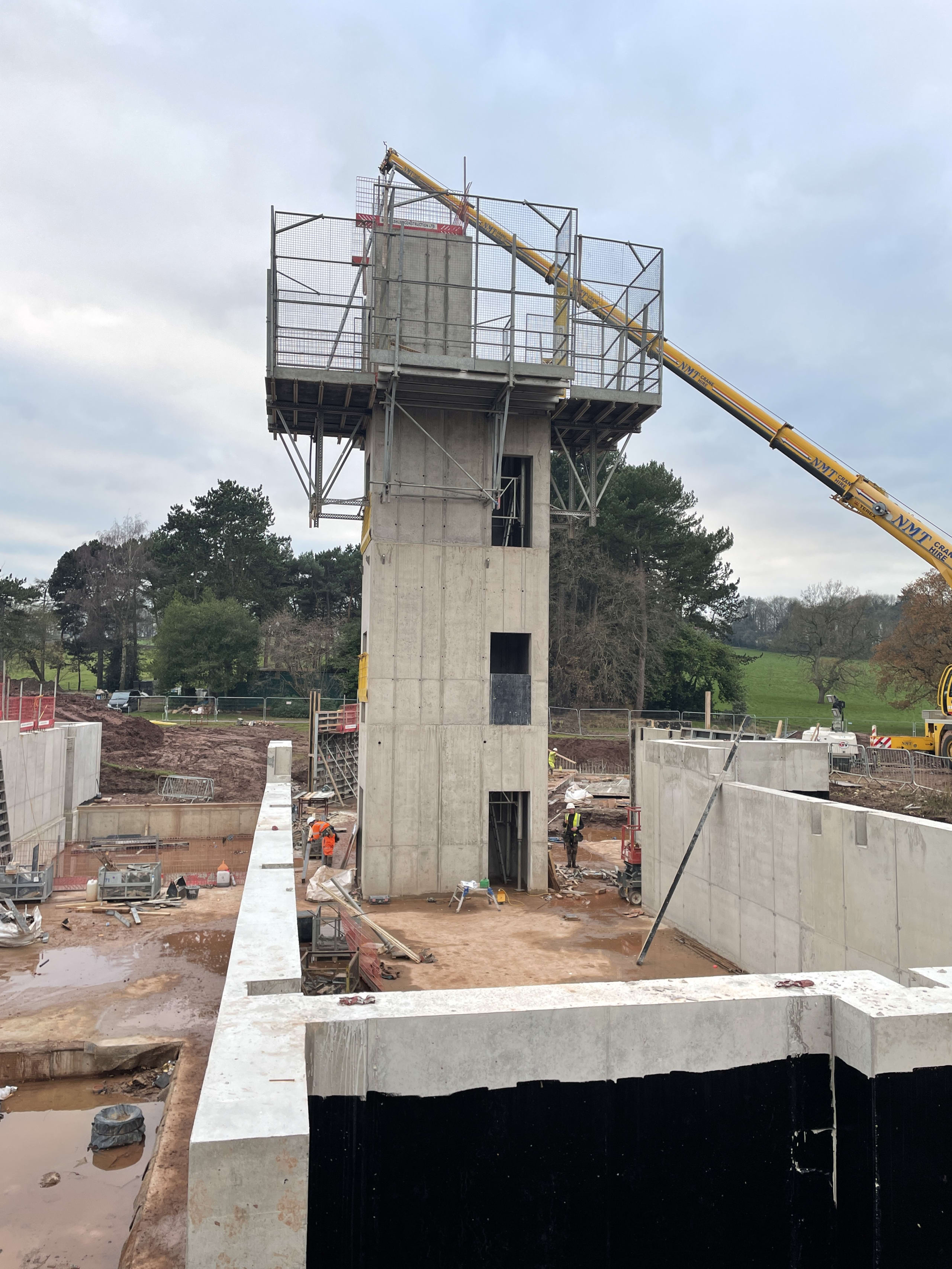 Concrete tower structure under construction with scaffolding platform, crane, and workers at water treatment facility site