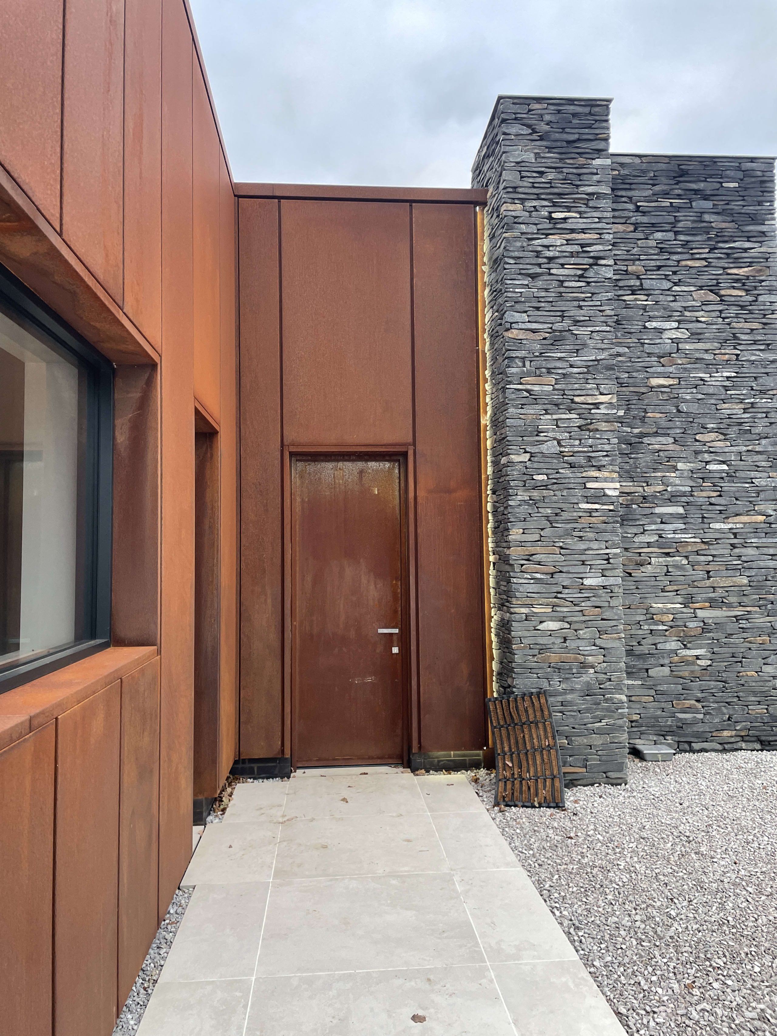 Modern entrance featuring weathered Corten steel cladding and door alongside dry stone wall with slate pathway