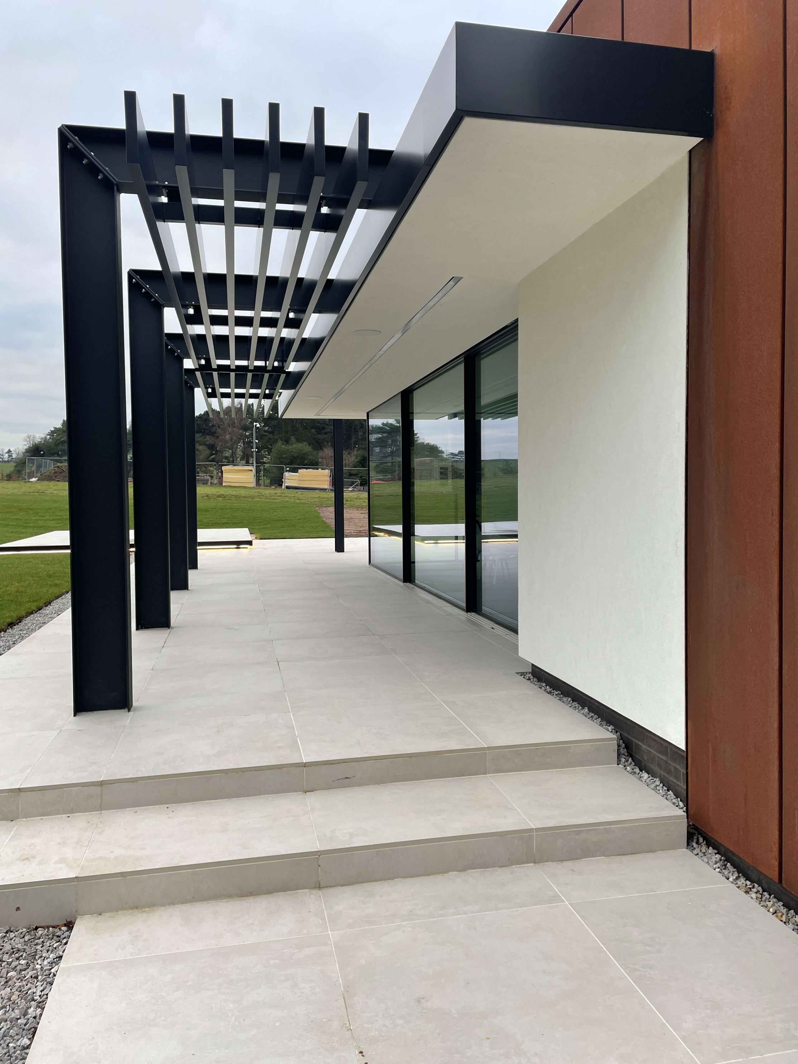 Modern building exterior with black steel pergola, corten steel cladding, large glass windows, and limestone patio tiles