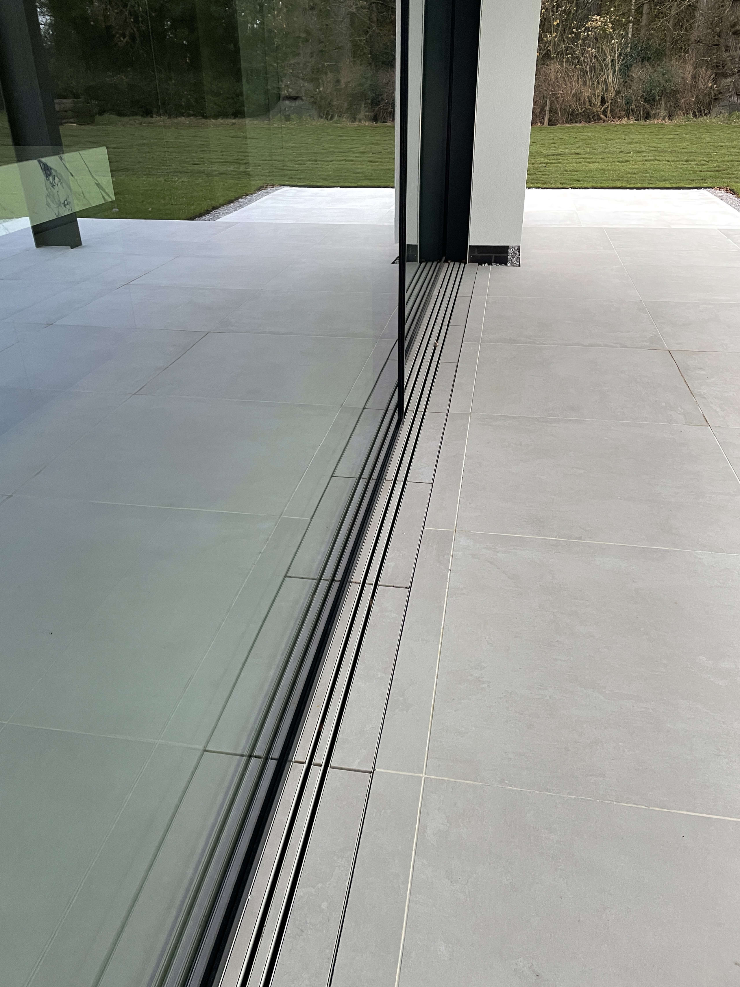Seamless indoor-outdoor porcelain tile flooring with recessed sliding door track and glass balustrade overlooking garden