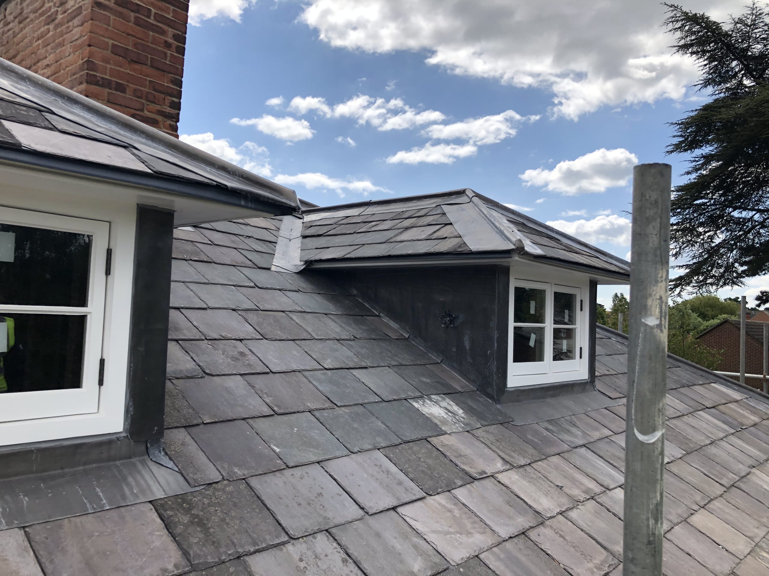 Slate-tiled roof with a white-framed dormer window and brick chimney under a partly cloudy sky