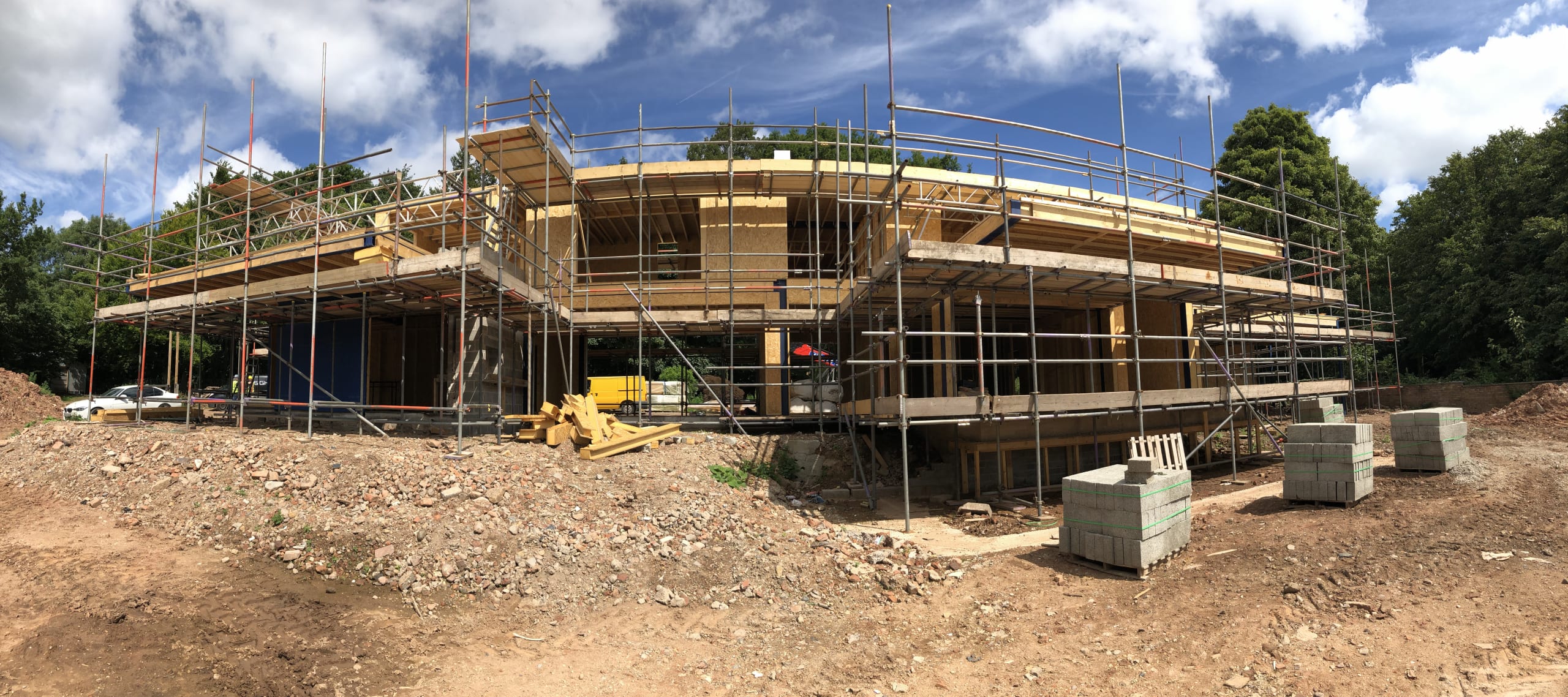 Timber-framed house under construction with full scaffolding wrap and curved roofline, surrounded by cleared earth site