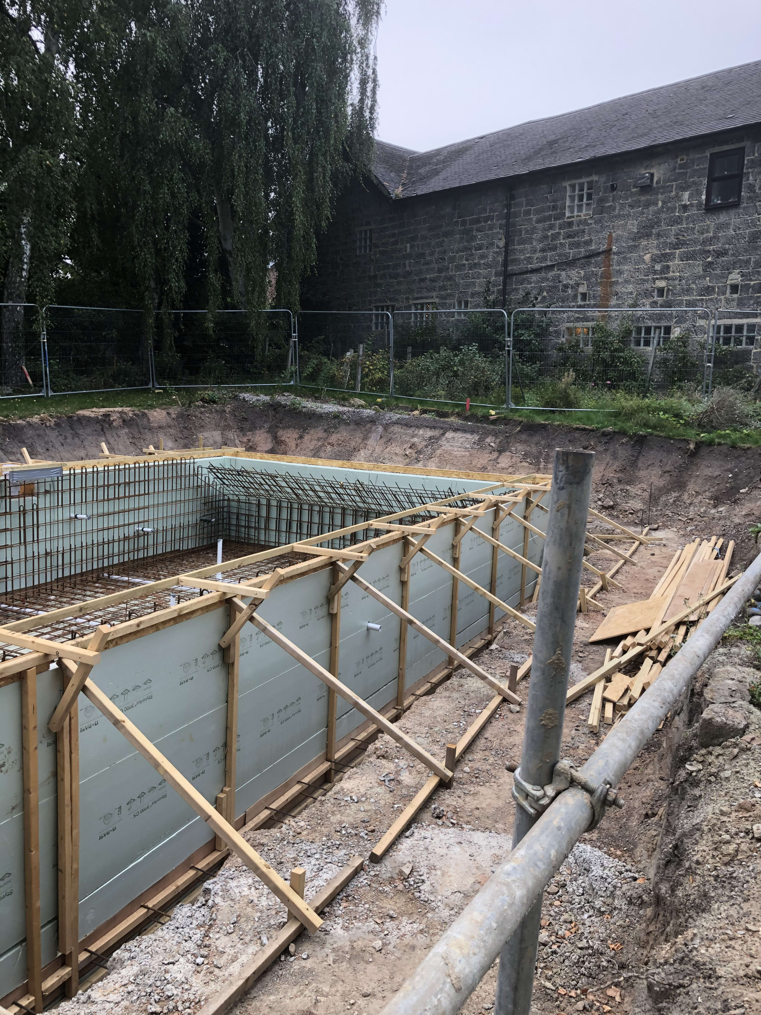 Excavated pool foundation with steel rebar grid and timber-braced formwork beside a stone barn