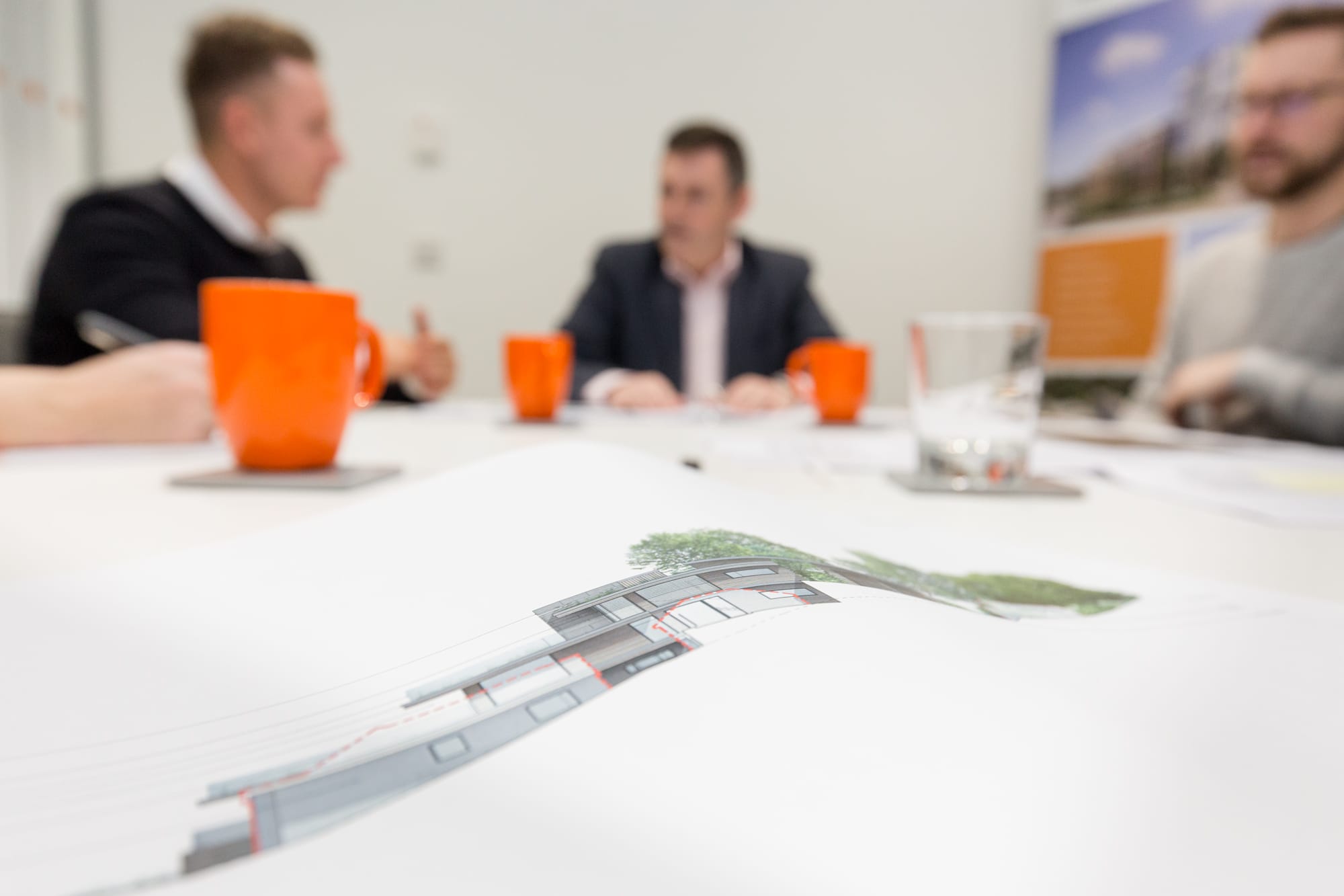 Architectural elevation drawing on a table during a design review meeting with three people discussing in the background