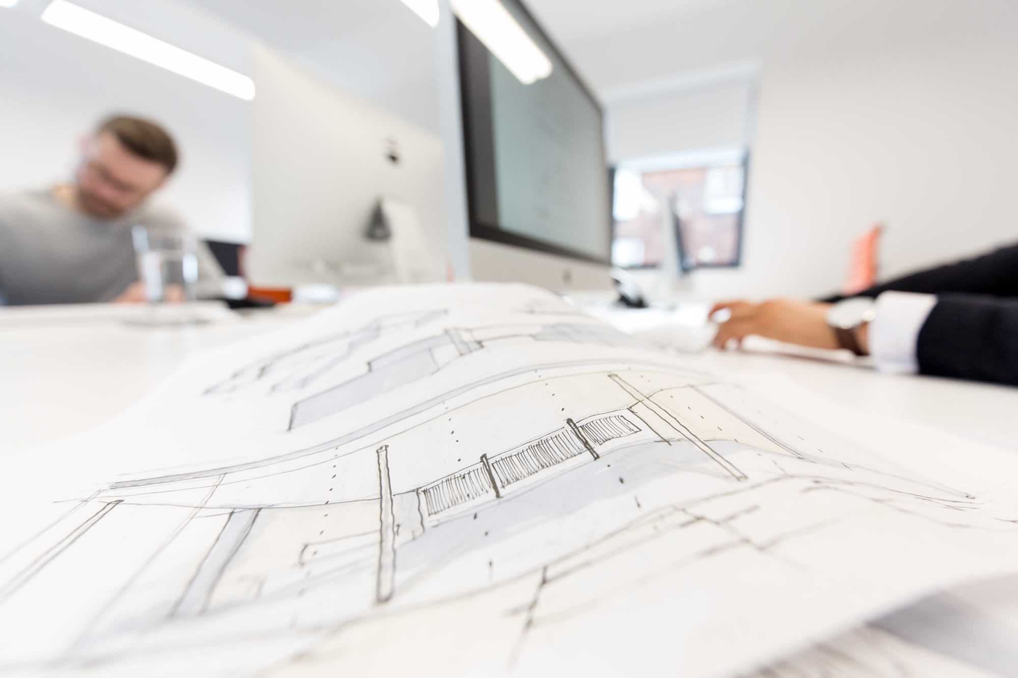 Hand-drawn architectural elevation sketch on a studio desk with designers working in the blurred background