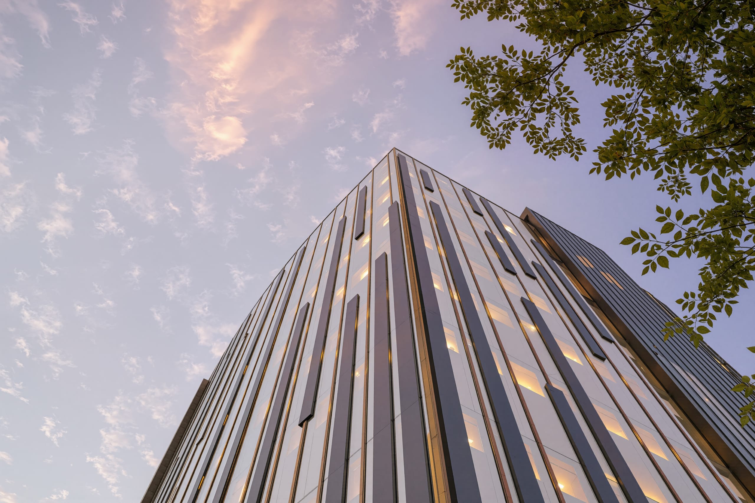 Modern high-rise building with vertical metallic facade panels photographed from ground level against twilight sky with trees