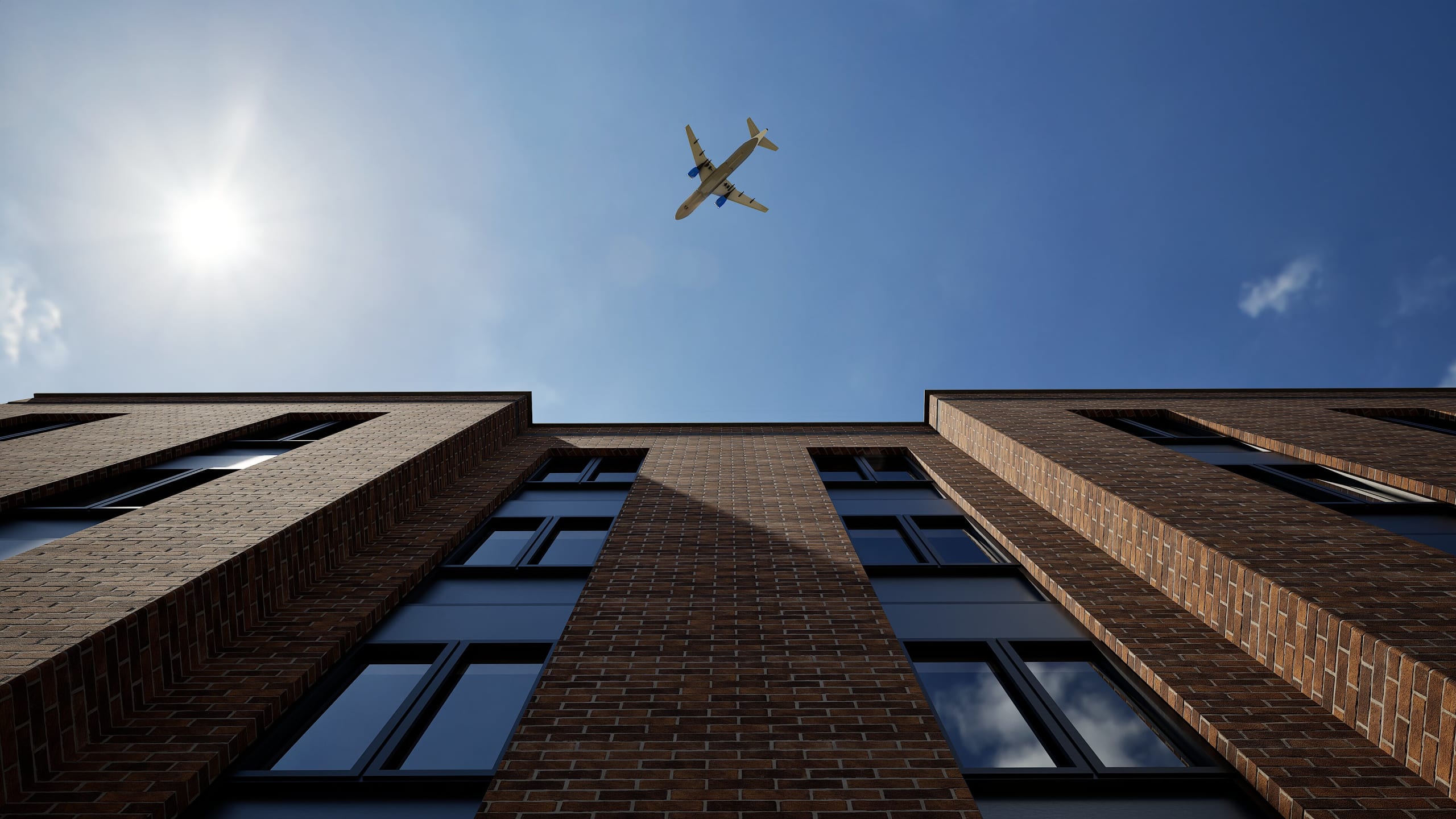 Upward view of modern brick apartment building with rectangular windows against blue sky with airplane flying overhead