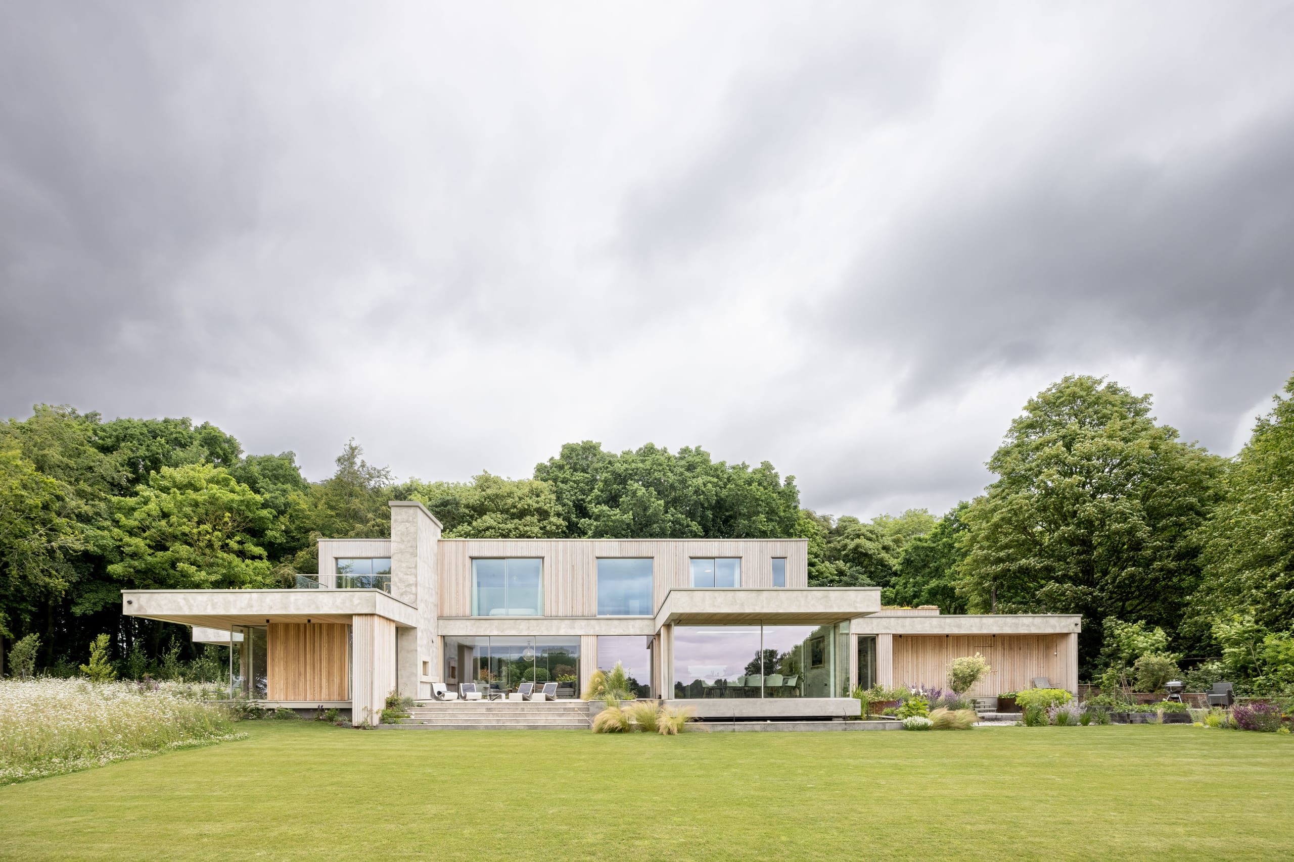 Contemporary two-story house with timber cladding and stone chimney overlooking a large lawn framed by mature trees
