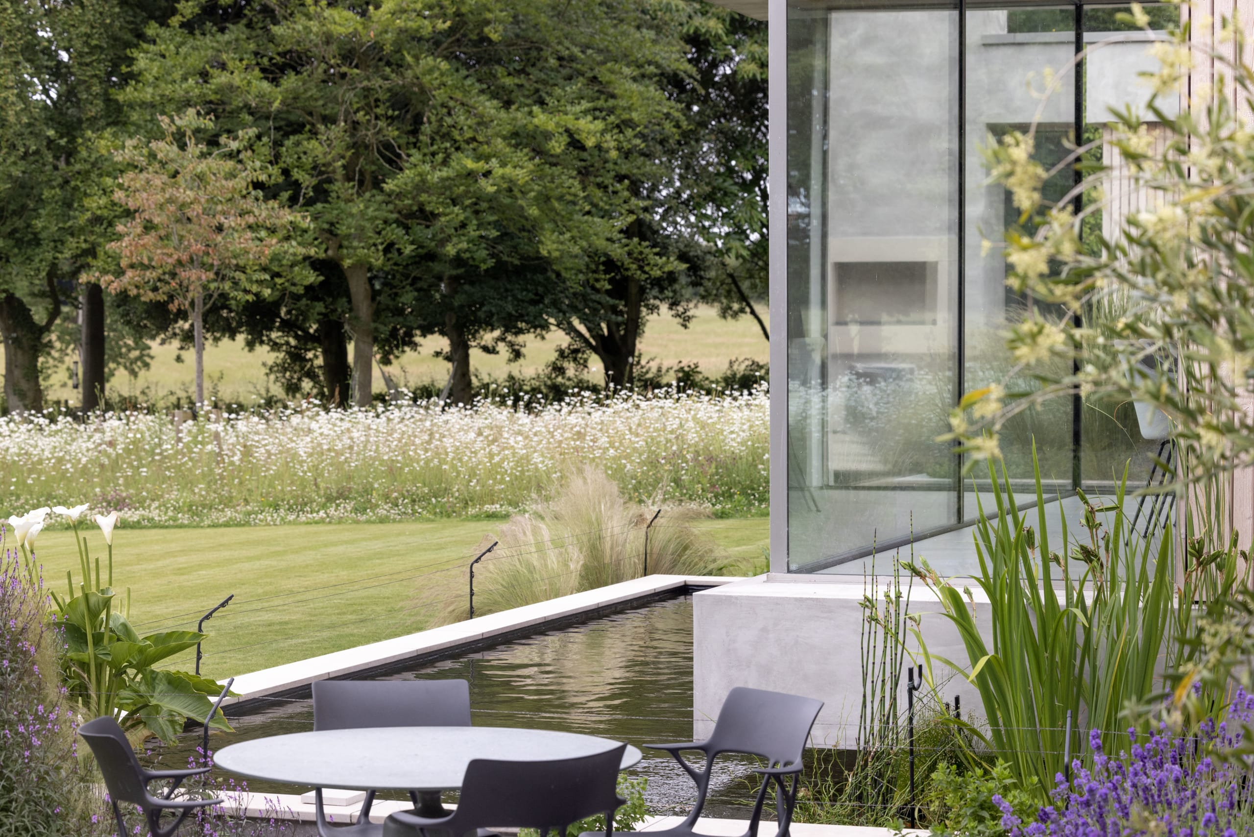 Glass-walled house corner overlooking a still reflecting pool with a wildflower meadow and lawn beyond