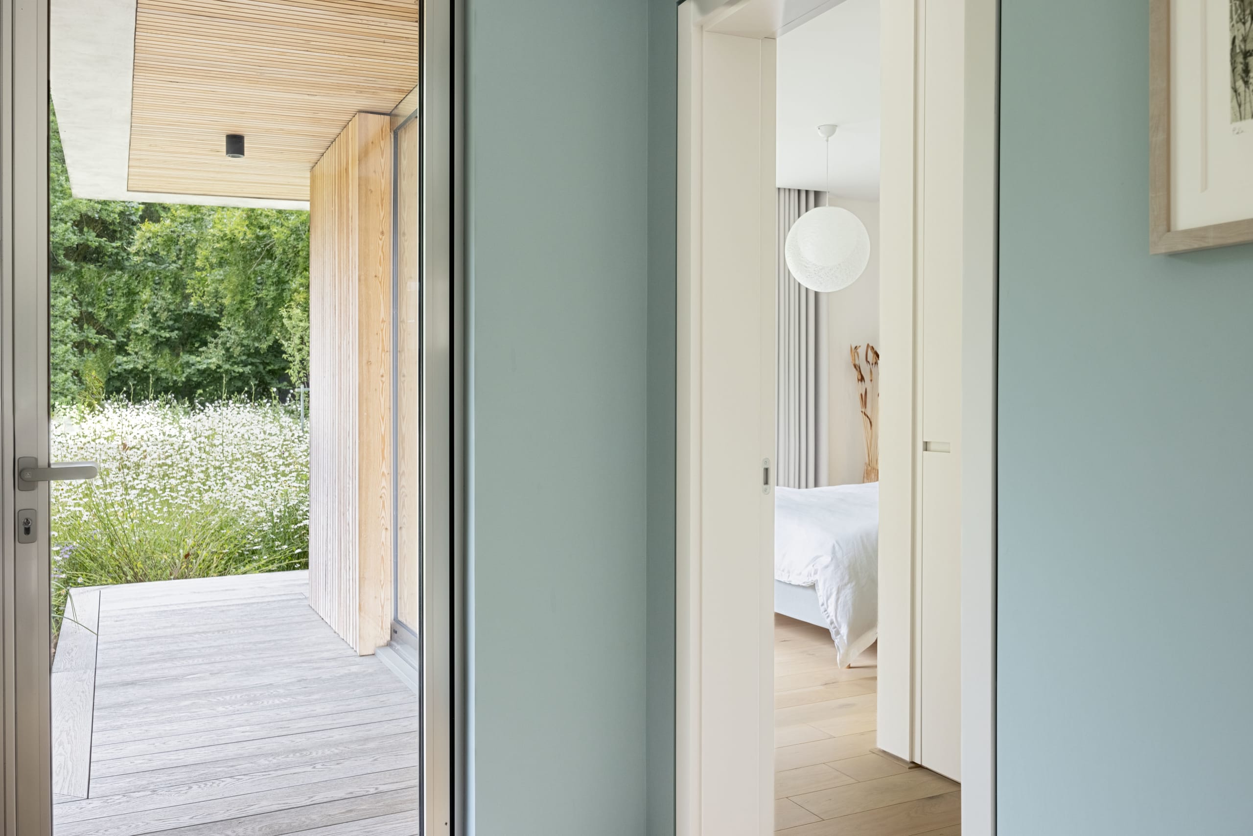 Split view of a timber-clad garden terrace and a soft blue-green hallway leading to a bedroom