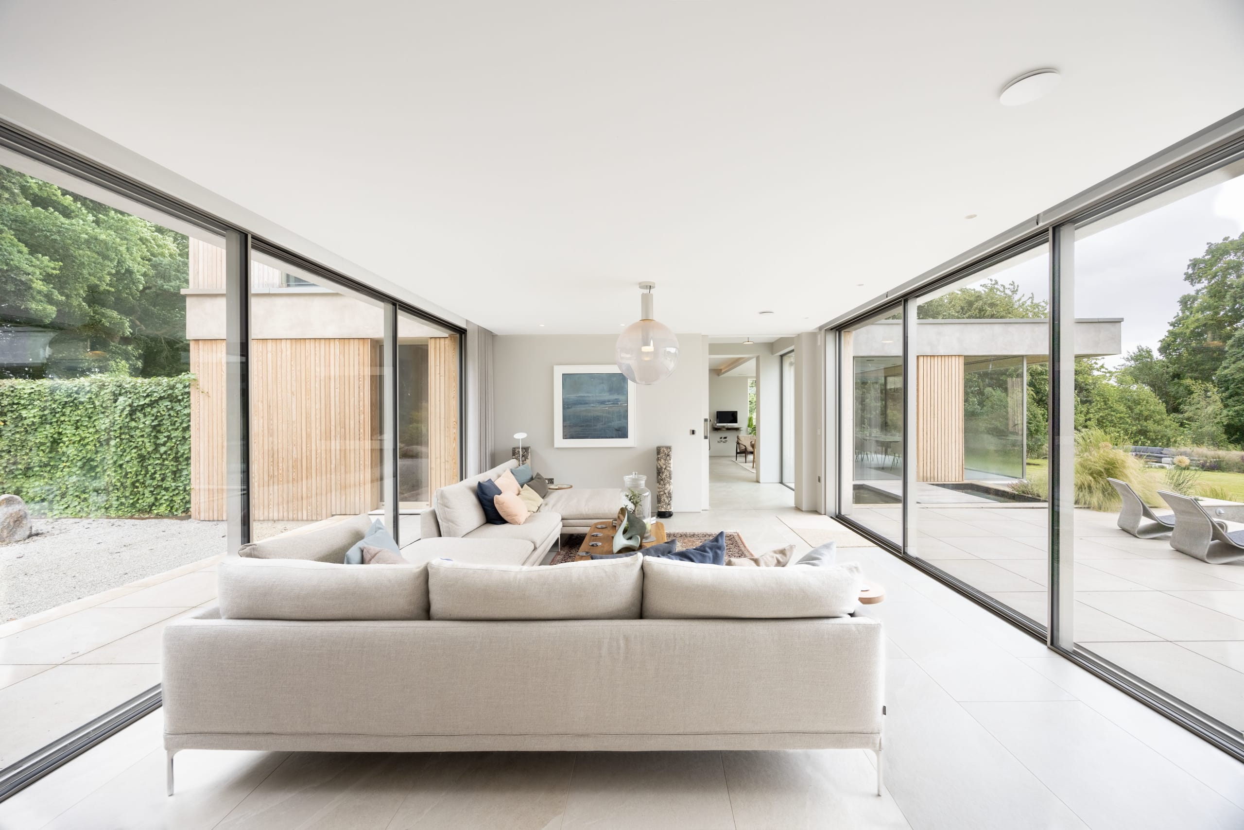 Living room with corner sofa framed by full-height glass walls opening to gardens on both sides