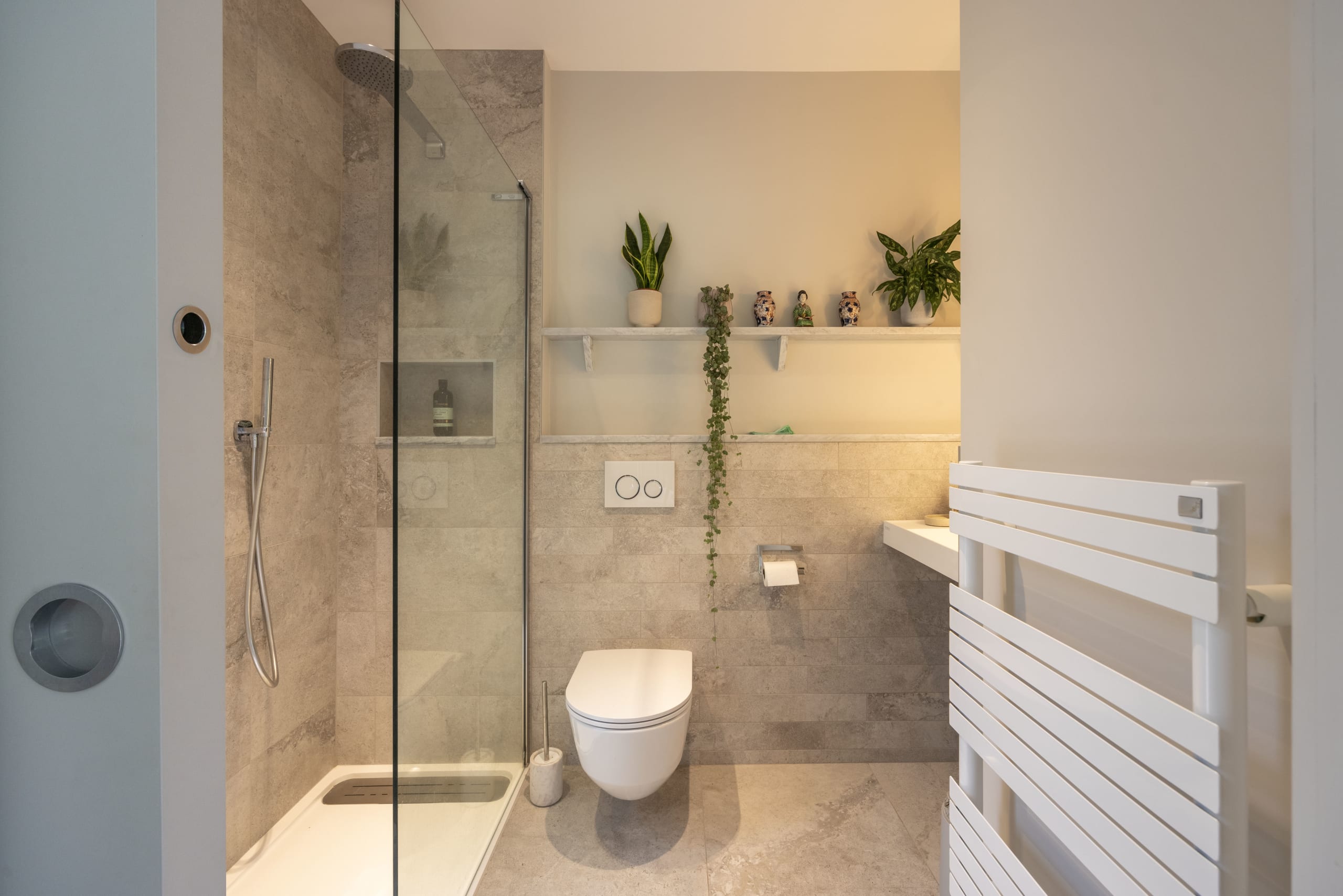 Bathroom with glass-enclosed shower, wall-mounted toilet, stone tile, and a white ladder towel radiator