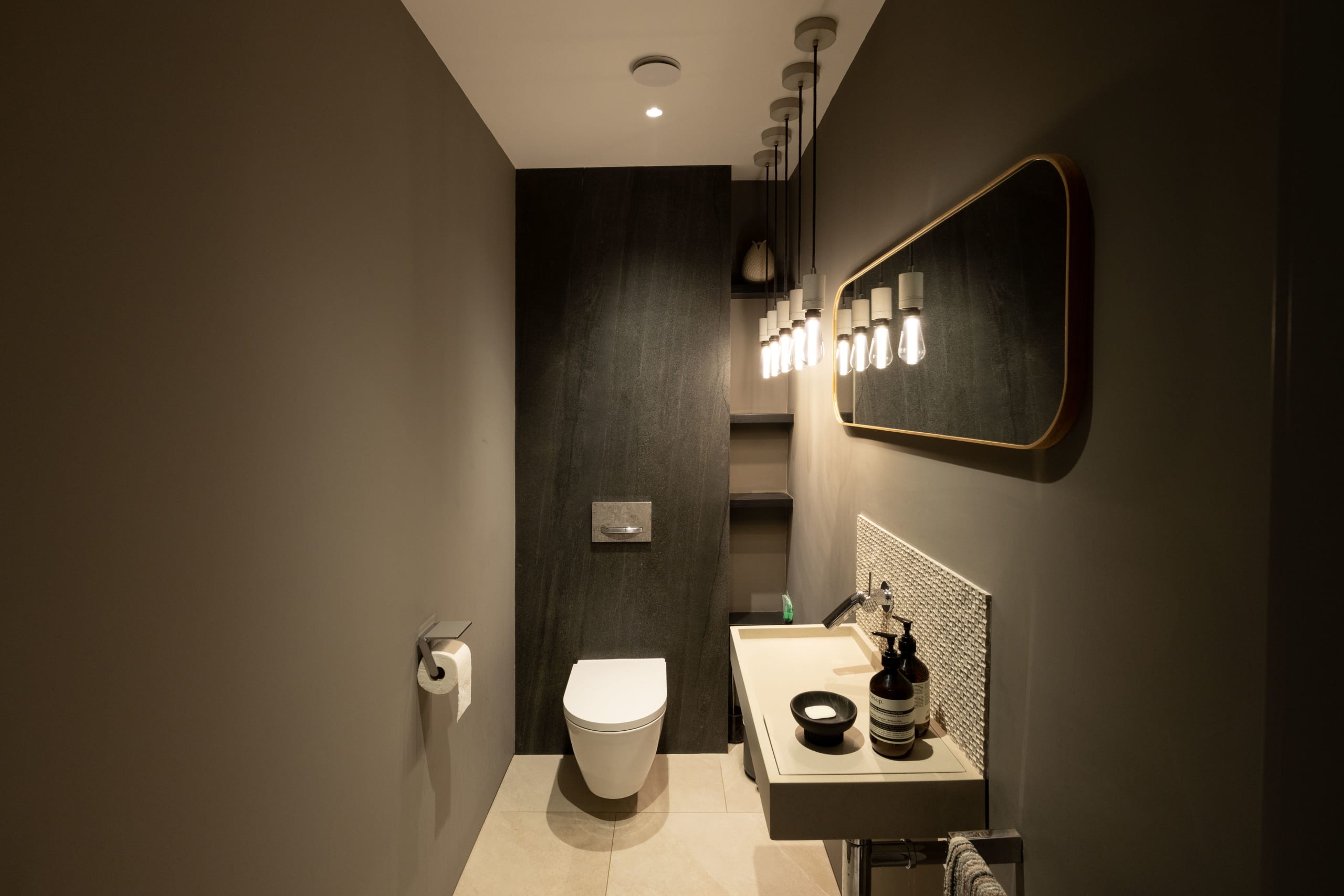 Dark powder room with wall-hung toilet, cluster pendant lights, and a floating vanity beneath an oval brass-framed mirror