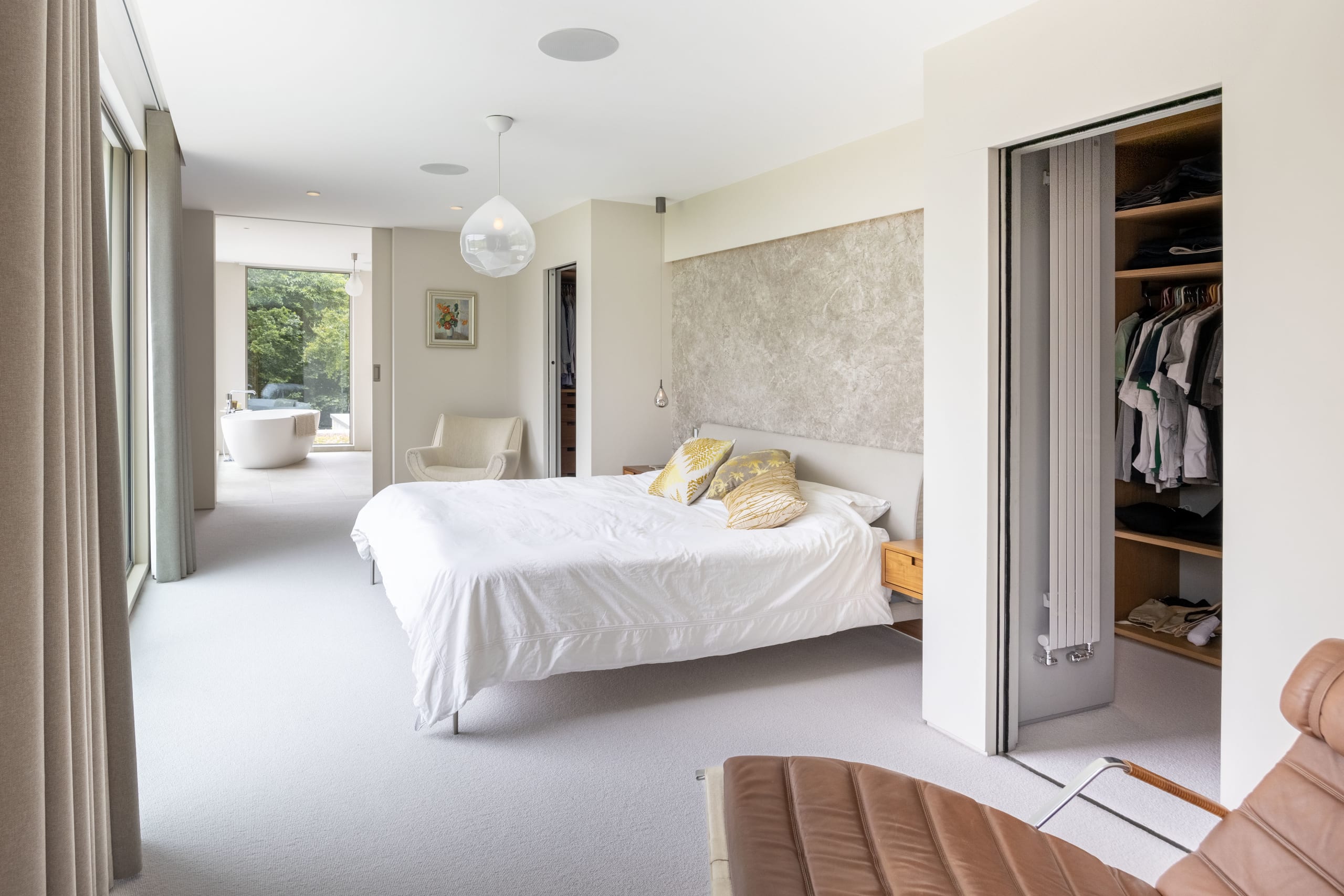 Bedroom suite with white bed, textured feature wall, open wardrobe, and a freestanding tub visible through an adjoining glazed bathroom