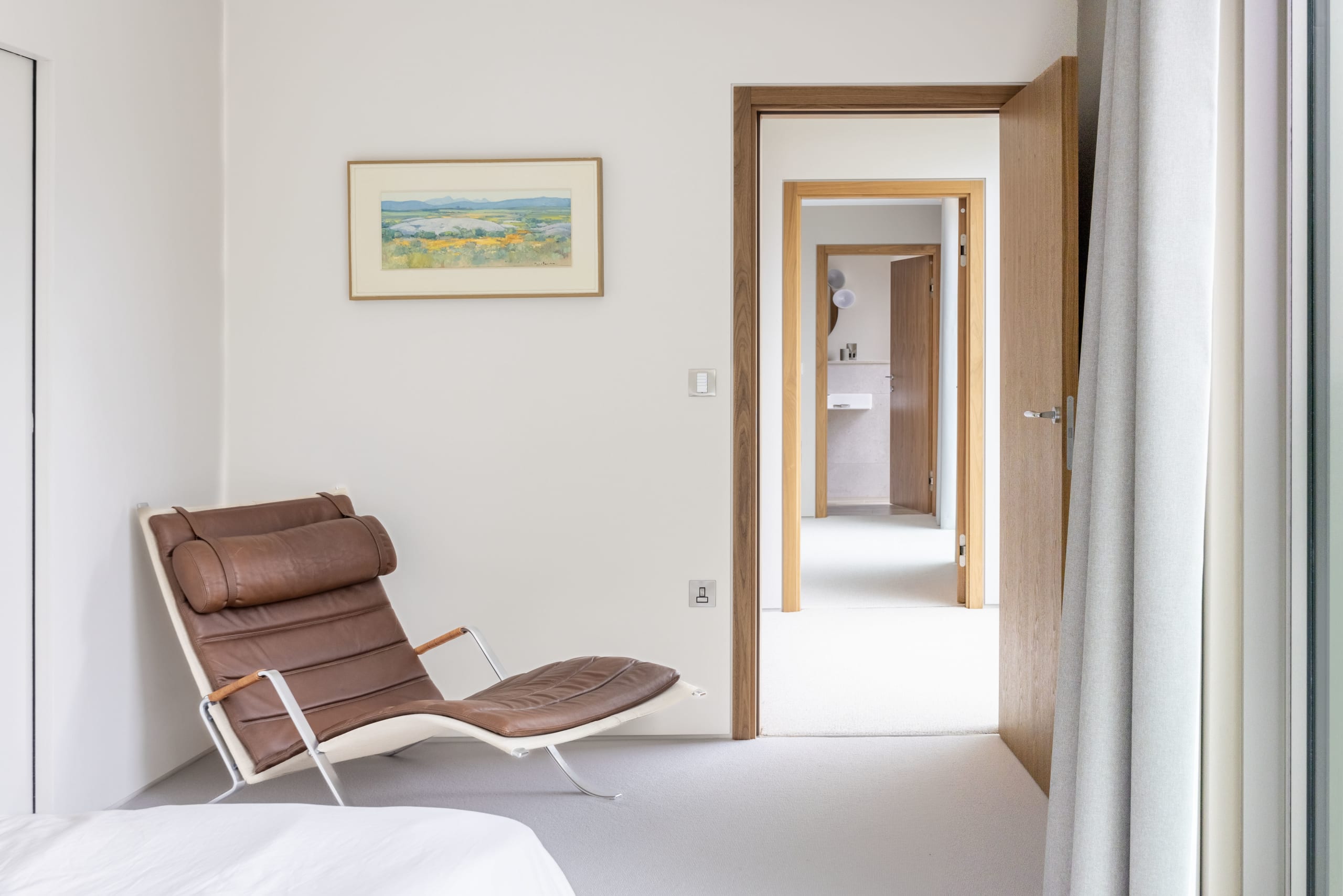 Bedroom corner with brown leather chaise lounge and framed landscape print beside an open door to an en-suite bathroom