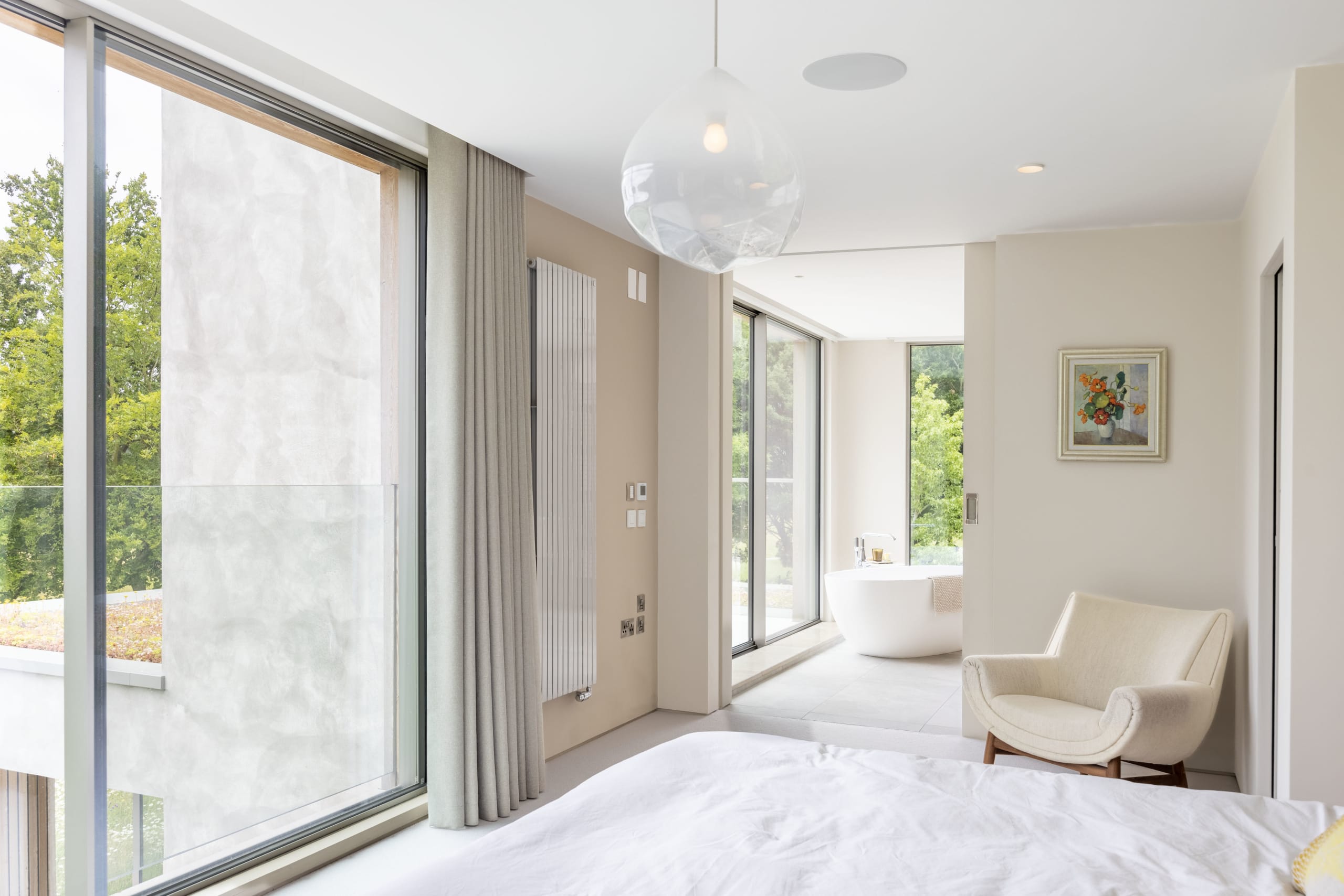 Bright bedroom suite with floor-to-ceiling windows, freestanding tub, and cream upholstered armchair