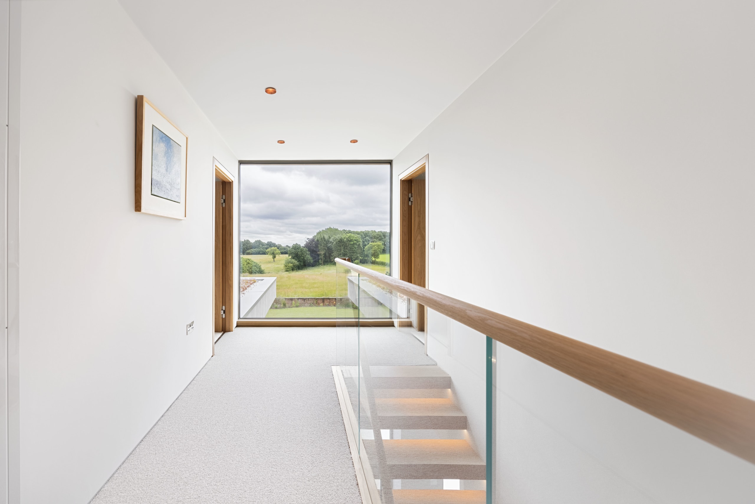 Upper-floor hallway with glass-and-timber stair balustrade framing a full-height window over green countryside