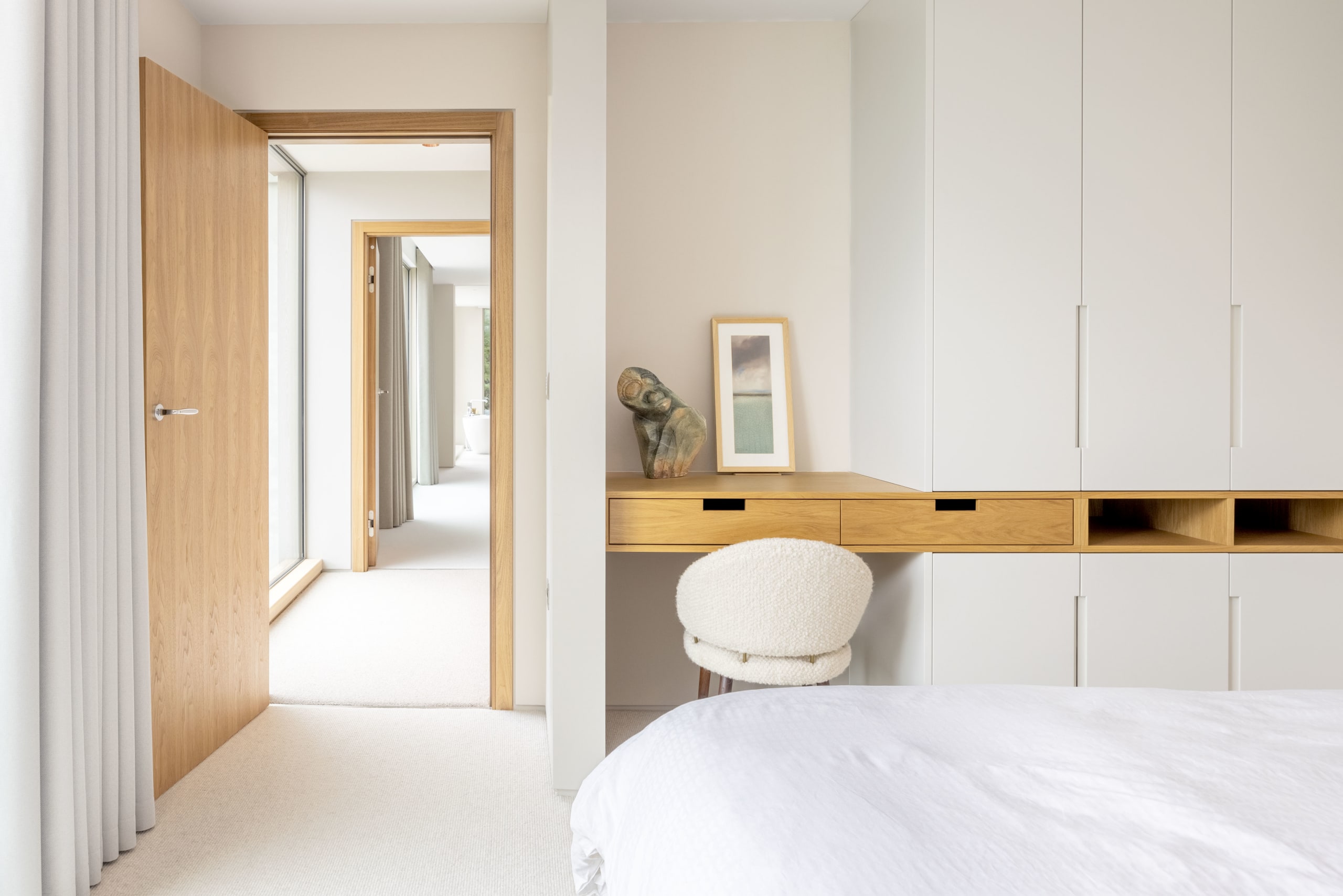 Minimalist bedroom with built-in oak desk, white wardrobes, and view through open door to hallway