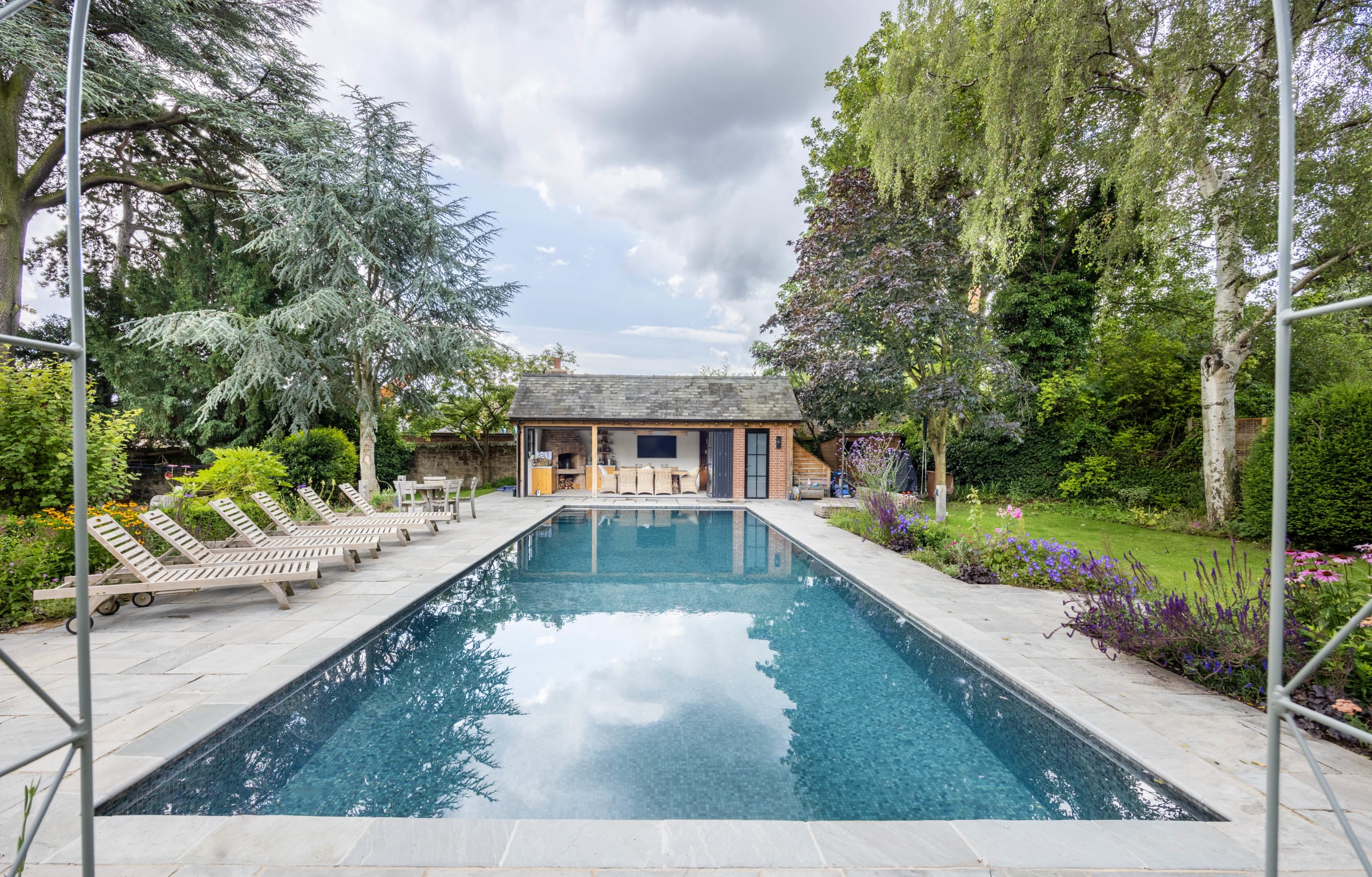 Rectangular garden swimming pool with stone coping leading to a timber-clad pool house